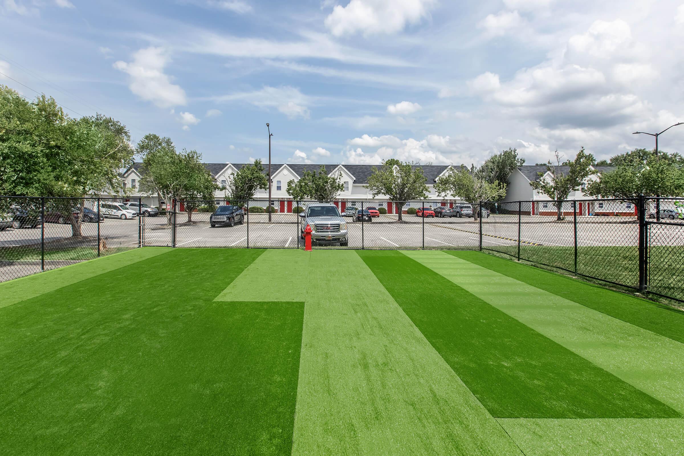 A fenced outdoor play area featuring green artificial turf. In the background, there are several parked cars and a residential building with multiple units. The sky is partly cloudy, creating a bright and inviting atmosphere.