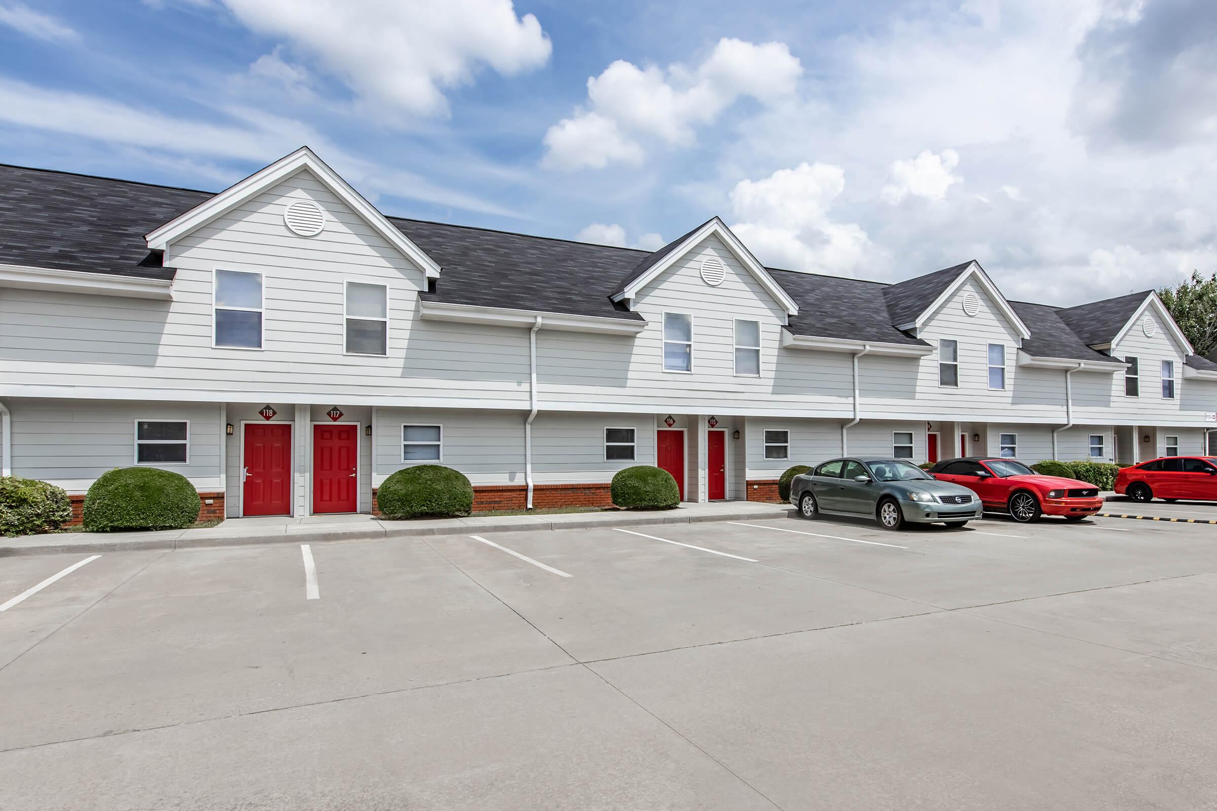 A row of white two-story residential townhomes with red front doors, situated in a parking lot. The area features well-manicured shrubs and a clear blue sky with scattered clouds. Cars are parked in front of the townhomes, with two clearly visible: a silver sedan and a red vehicle.