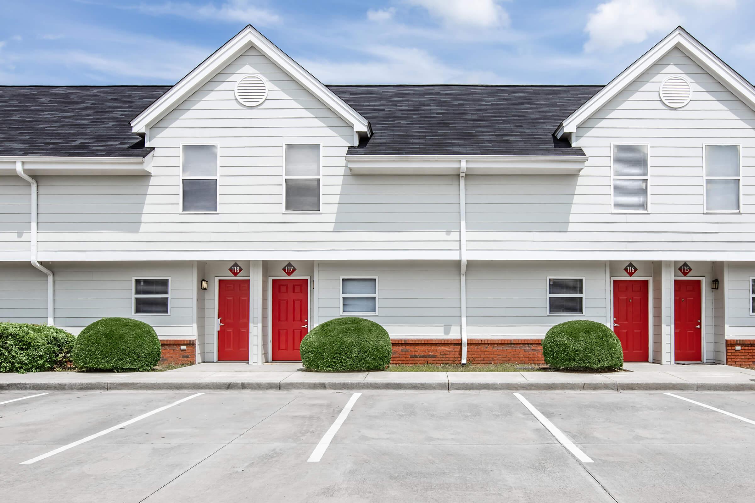 A row of two-story townhouses with a light gray exterior and red front doors. The building features a symmetrical design, with two doorways visible. Neatly trimmed bushes are planted in front, and a concrete parking area is empty, with several parking spaces marked. The sky is partly cloudy.