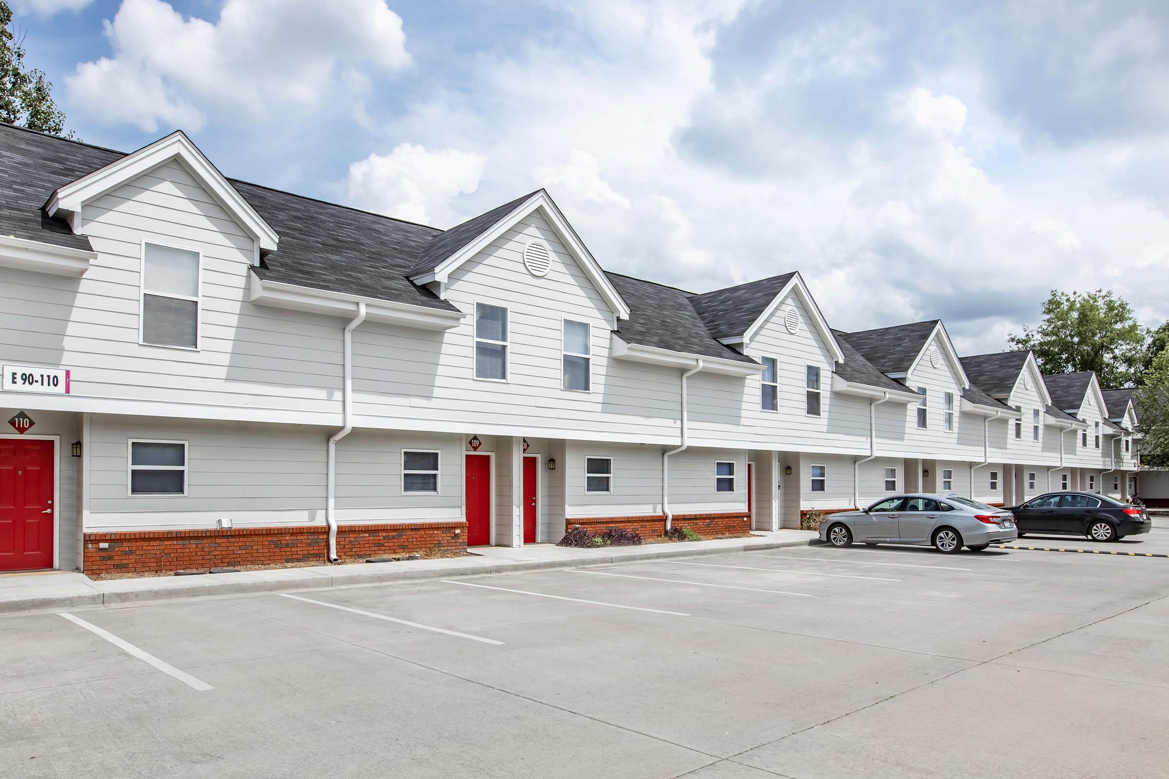 A row of two-story residential buildings with white siding and dark roofs. Each unit has red doors, and the area in front features a concrete parking lot with two parked cars. The sky is partly cloudy, adding a bright atmosphere to the scene.
