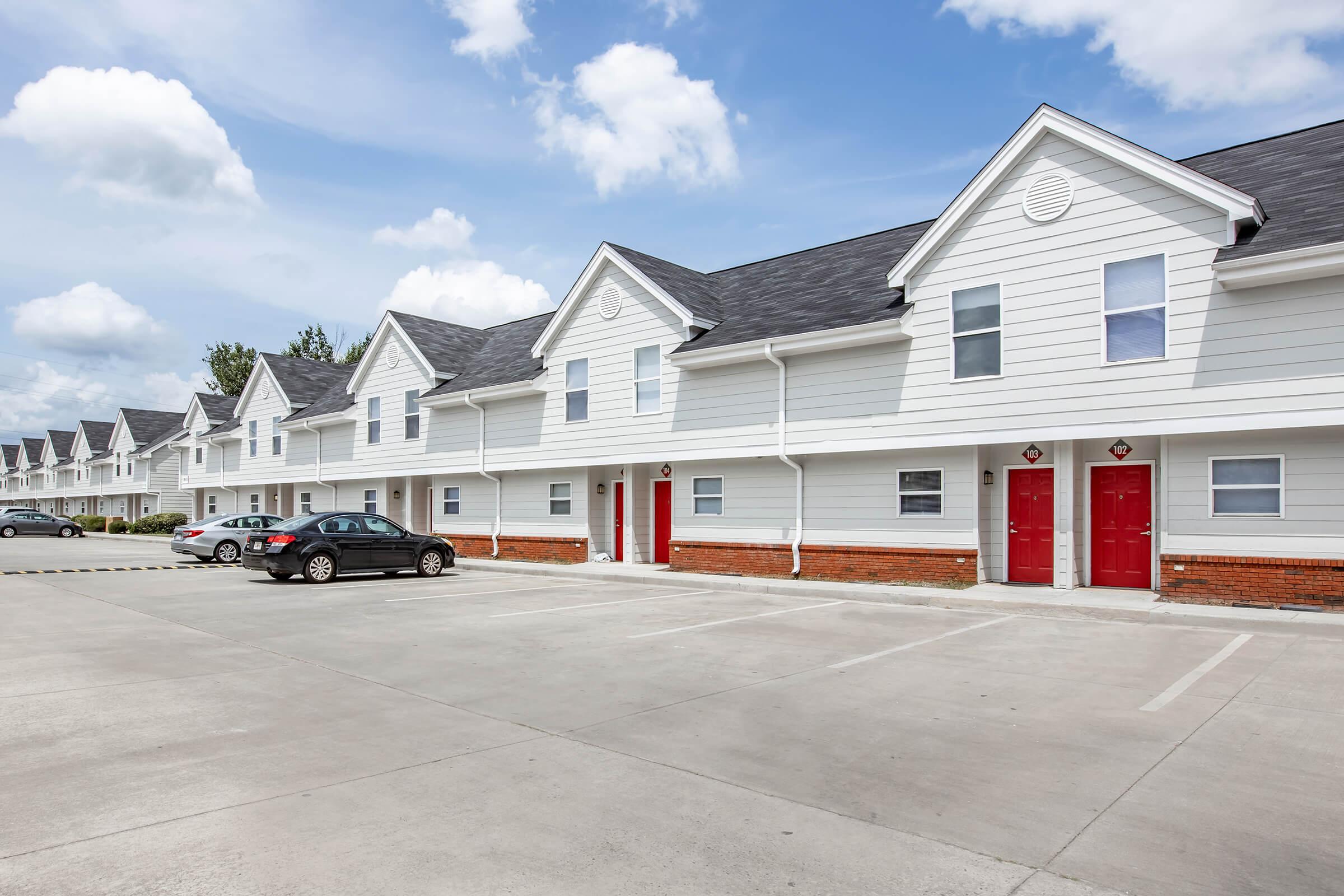 A row of white multi-story townhouses with peaked roofs and red doors. Clear blue sky with scattered clouds above. A concrete parking lot in front of the buildings, with parked cars. The scene suggests a residential area or apartment complex.