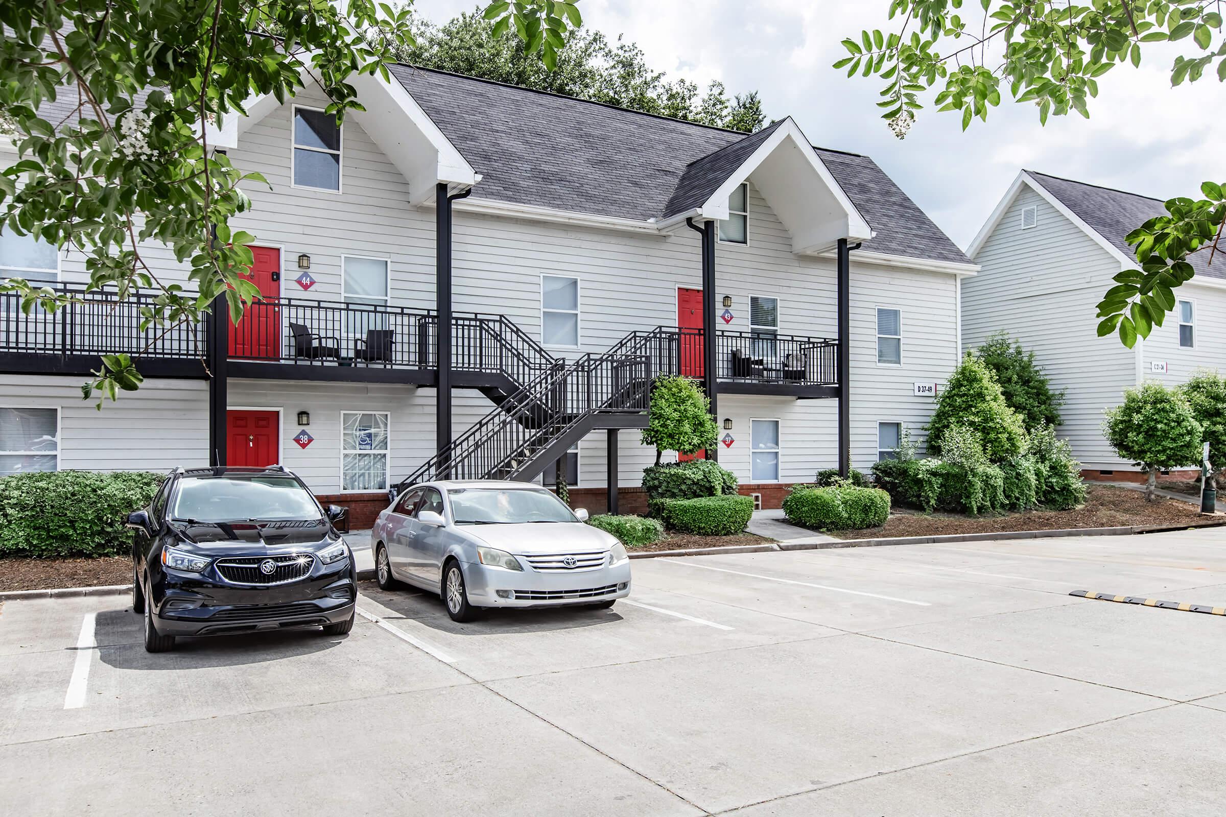Two parked cars in a parking lot in front of a two-story apartment building. The building features a light-colored exterior with red doors and a black staircase. Lush green shrubs and trees are visible around the entrance, and the sky is partly cloudy.