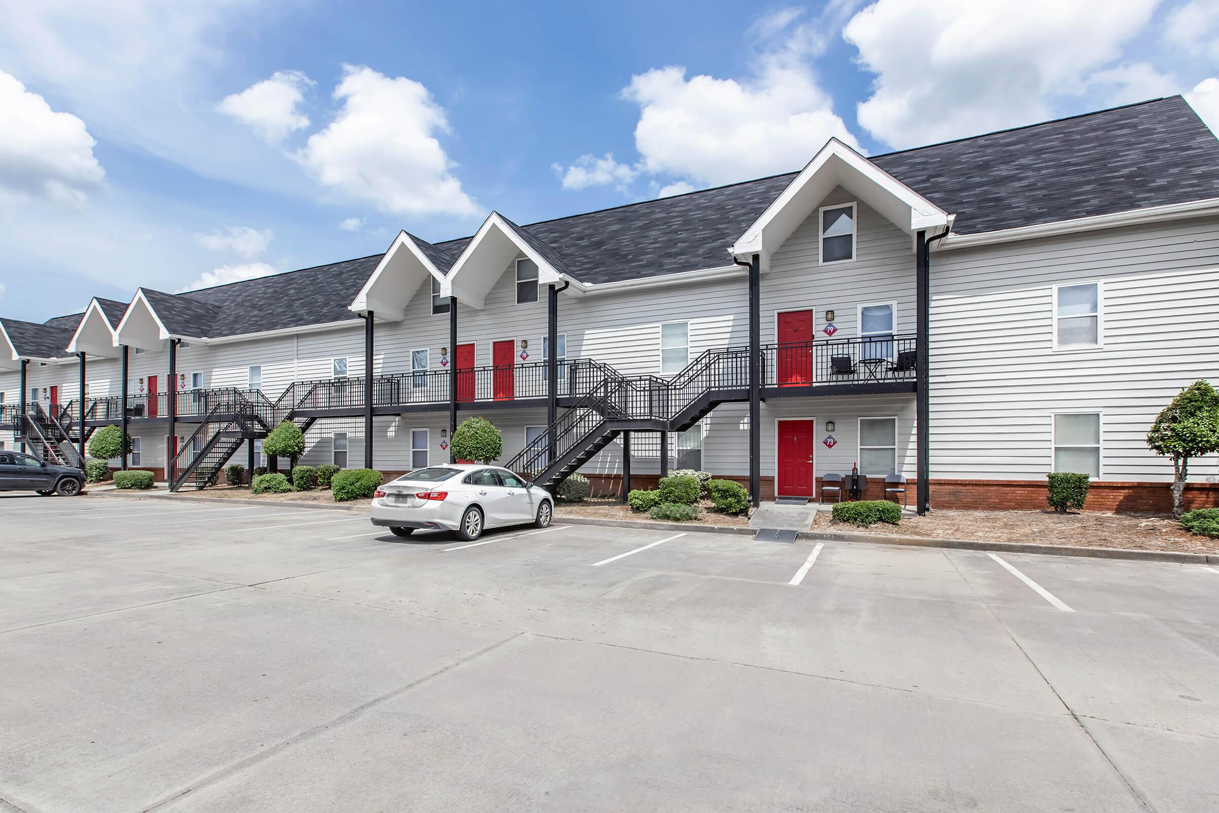A modern apartment complex featuring multiple two-story buildings with white siding and red doors. The scene includes a parking area in front, landscaped with small bushes, under a partly cloudy blue sky.