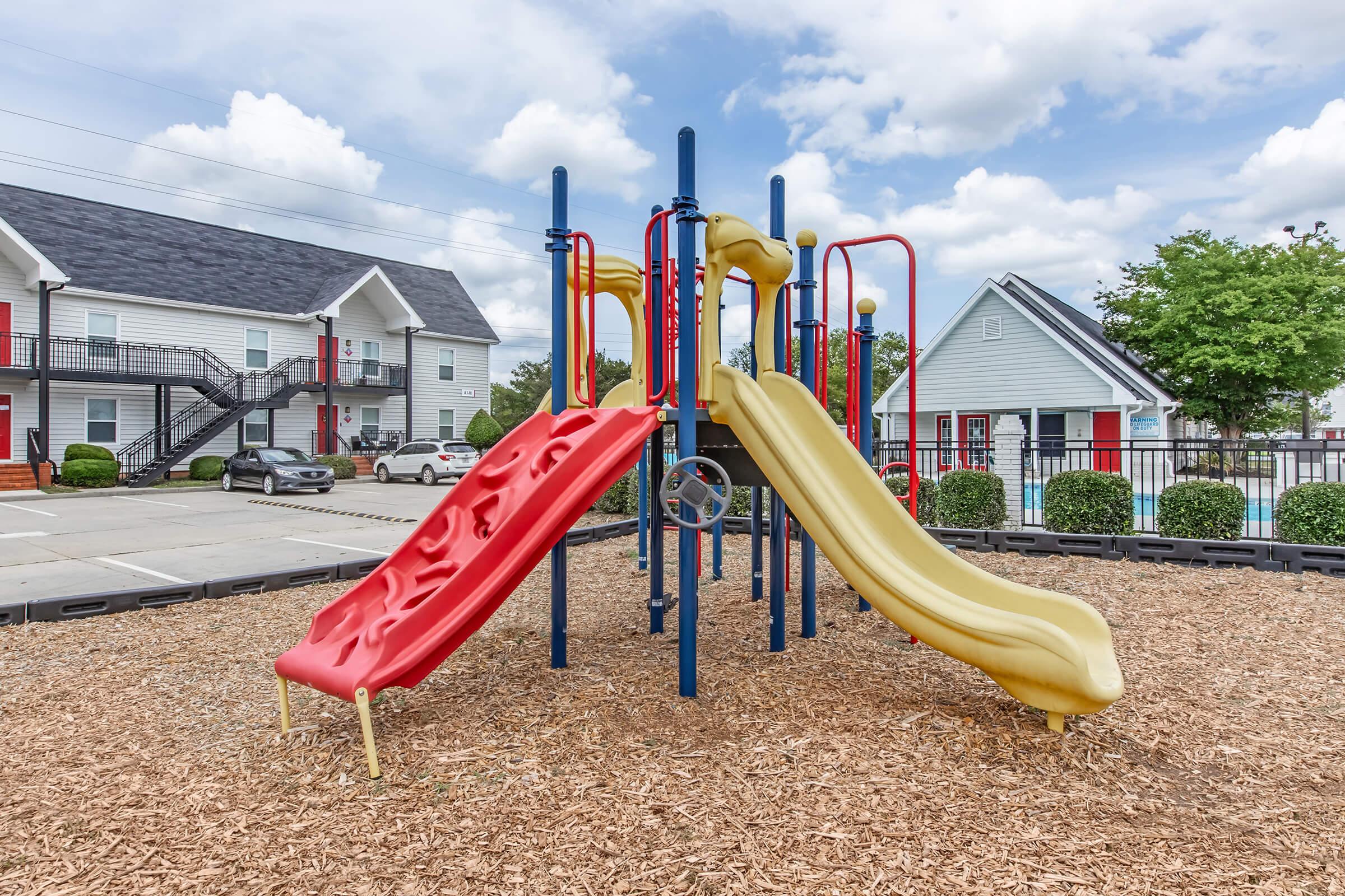 Colorful playground equipment featuring two slides, one red and one yellow, situated on a bed of wood chips. The background includes residential buildings and neatly trimmed bushes under a partly cloudy sky.