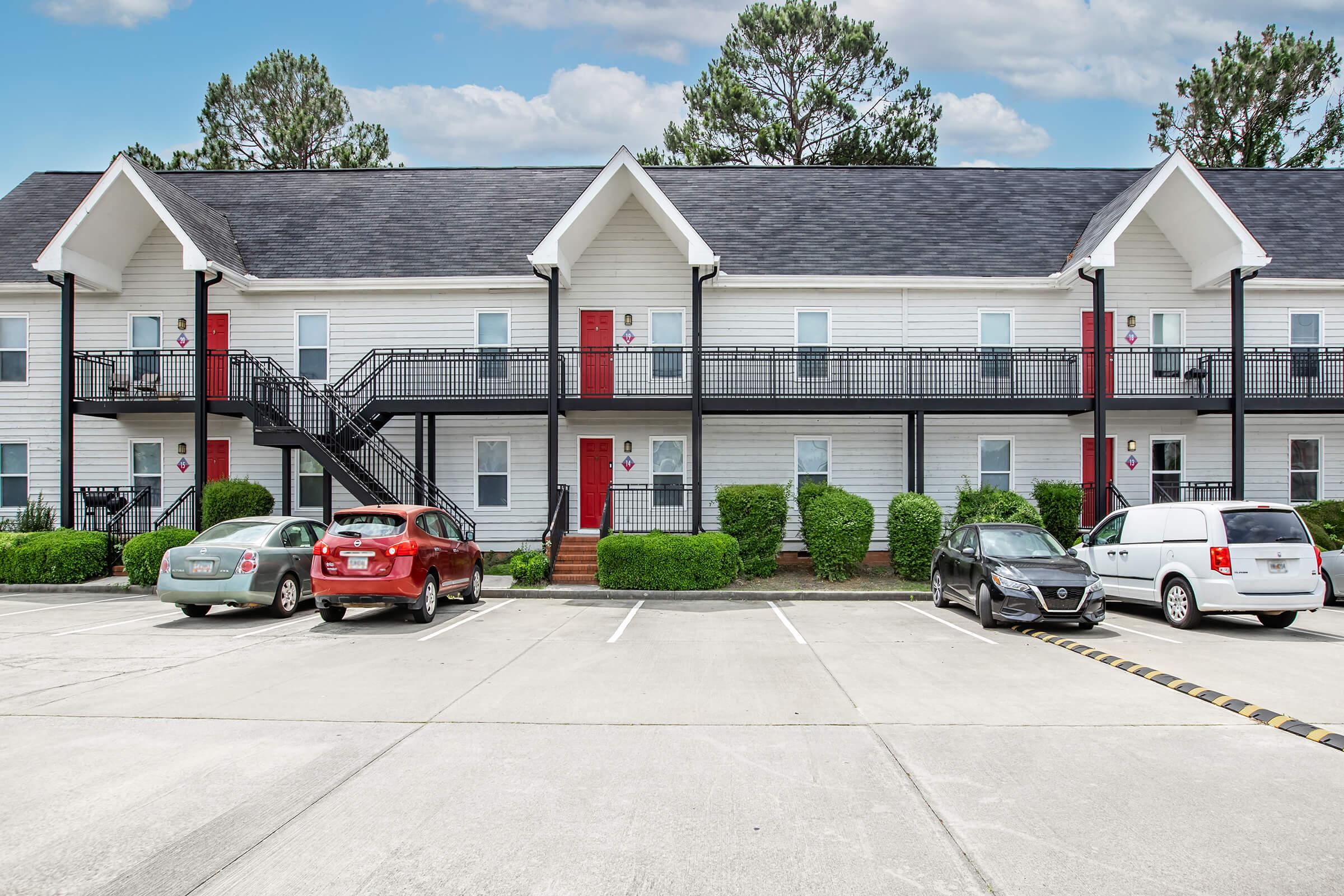 A modern two-story apartment building with a white facade and red doors. There are multiple parked cars in front, and well-maintained greenery surrounds the area. The building features black railings and a staircase, set against a blue sky with clouds.