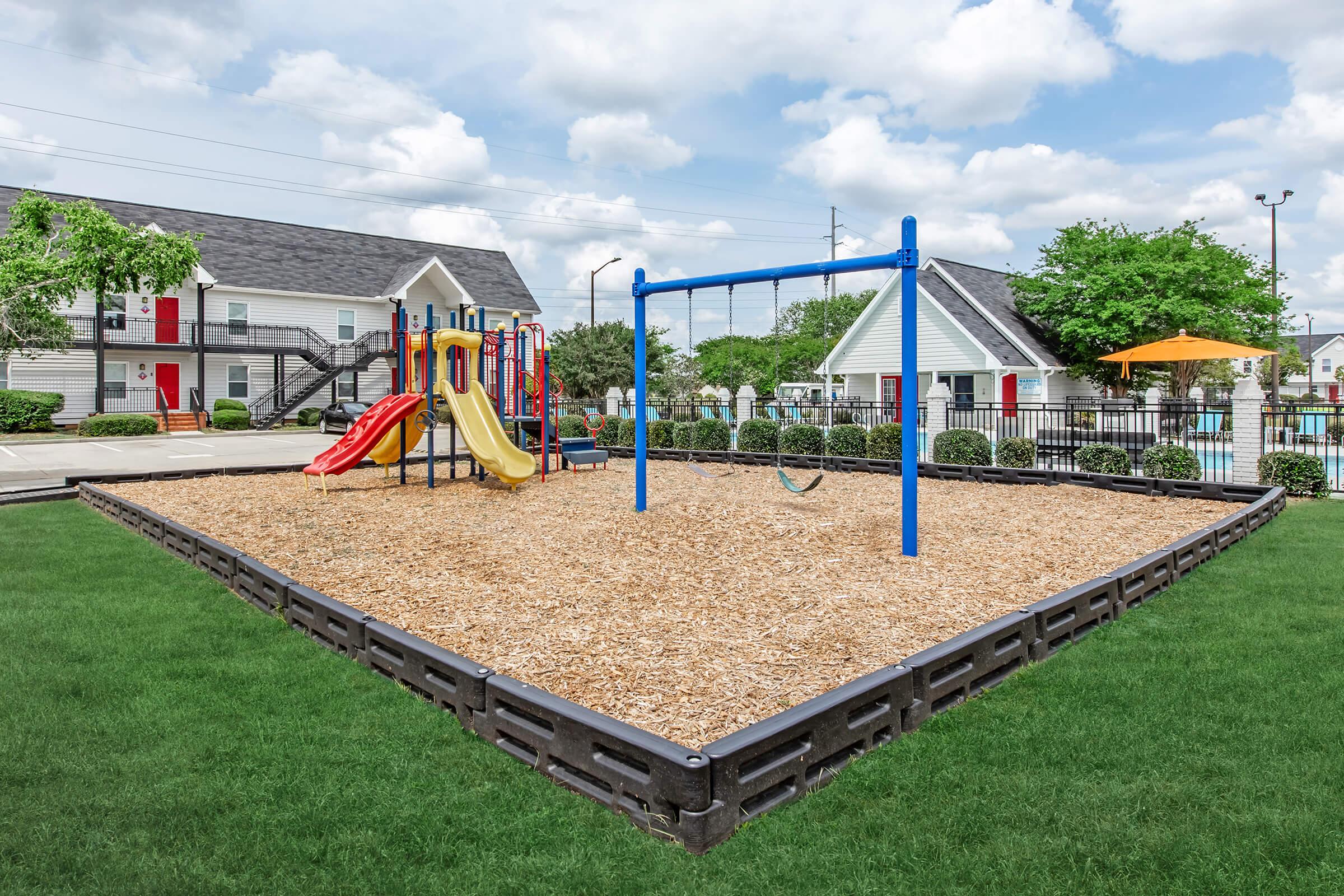 A colorful playground featuring a slide and swings, surrounded by wood chips and bordered by a black safety border. The playground is situated in a grassy area near residential buildings, with a clear blue sky and fluffy clouds in the background.