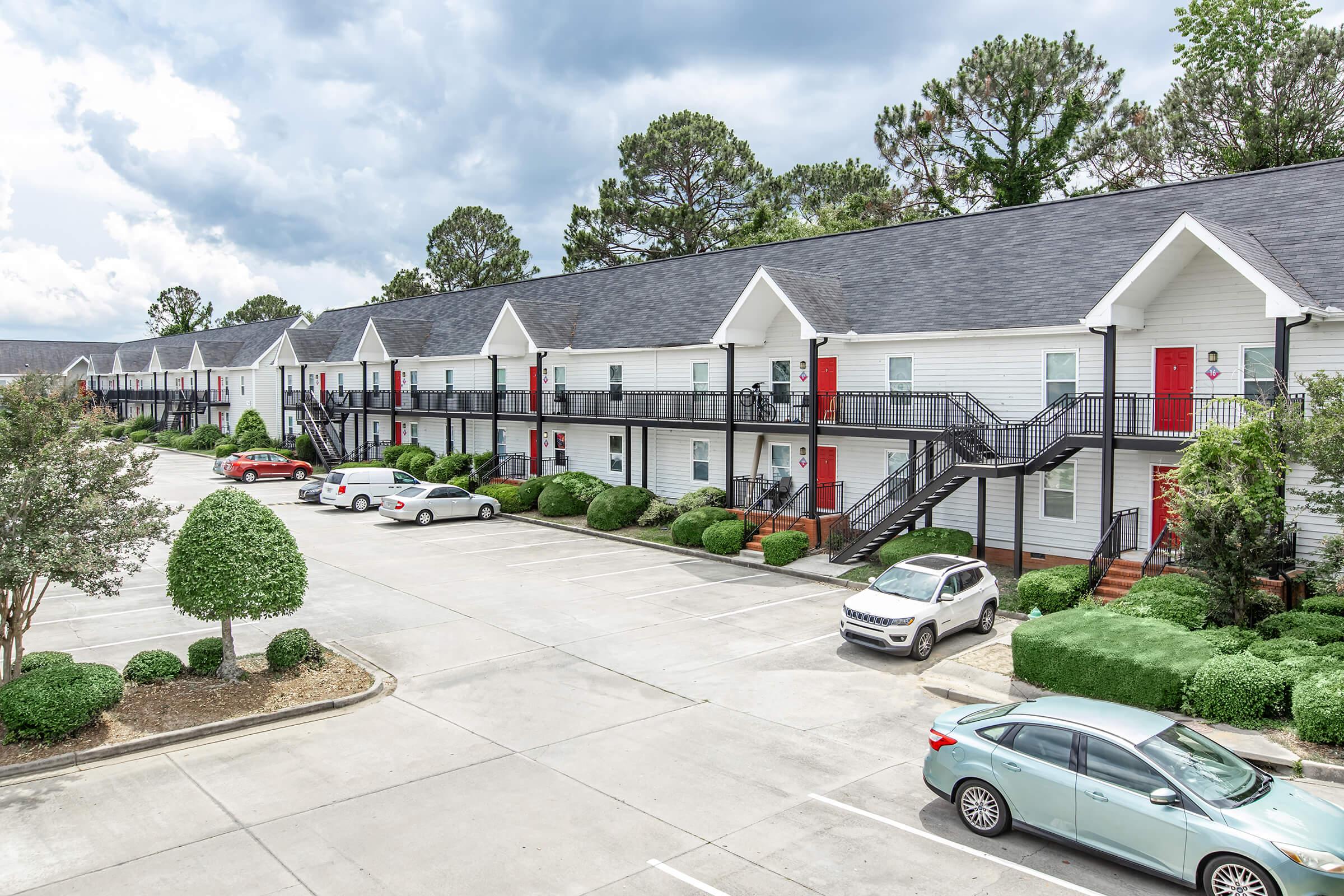 Row of apartment buildings with white exteriors and red doors, surrounded by neatly trimmed bushes and trees. A parking lot is visible in front, with several vehicles parked. The sky is partly cloudy, adding to the overall serene atmosphere of the residential area.