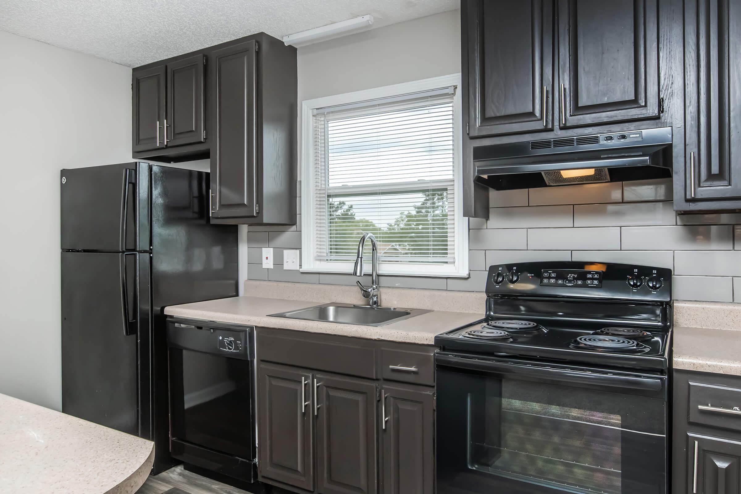 A modern kitchen featuring dark wooden cabinets, a black refrigerator, a black stove, and a stainless steel sink. The countertops are light-colored and there is a window above the sink, allowing natural light to enter the space. The flooring is a neutral color, complementing the kitchen design.
