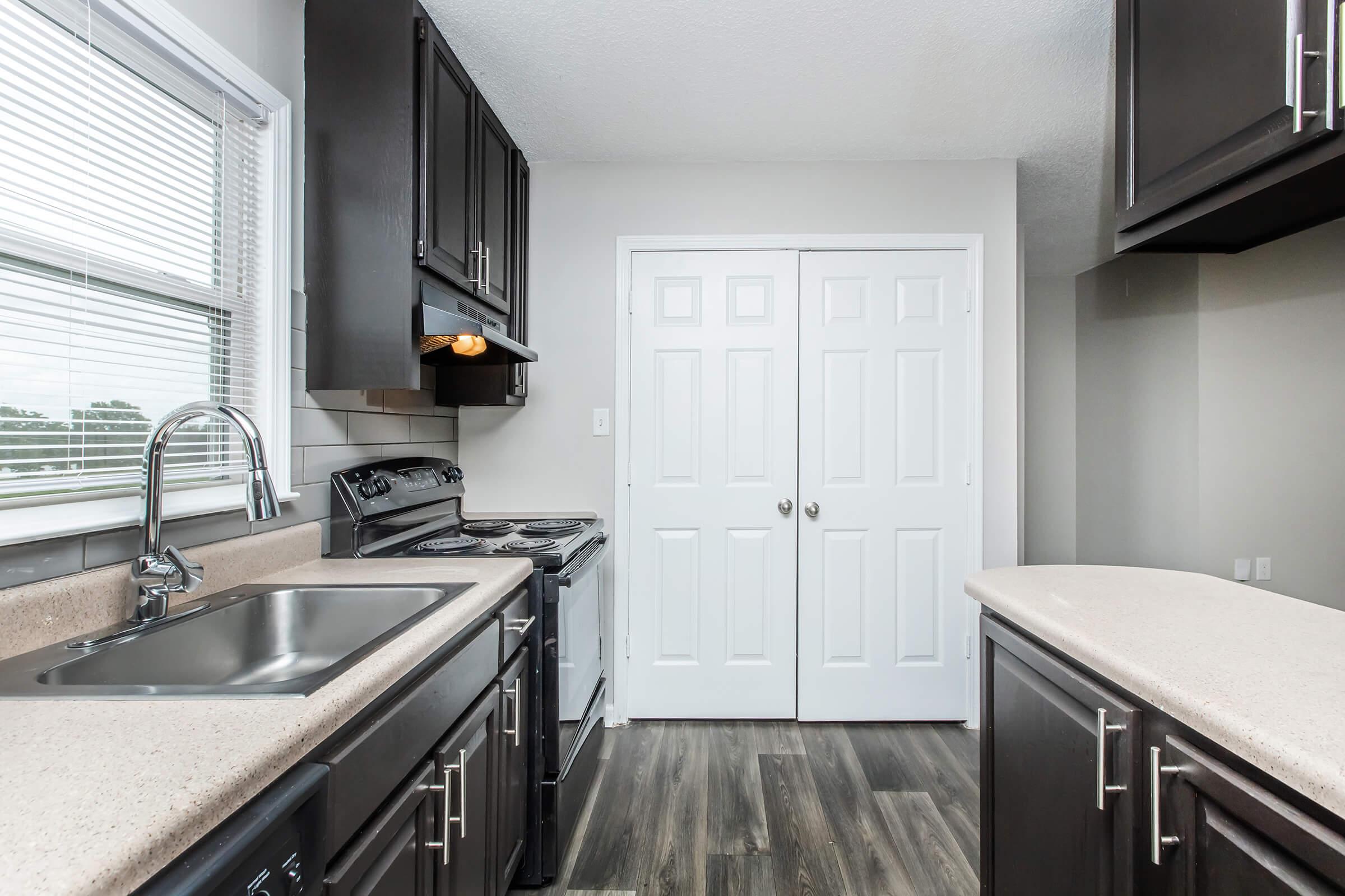 Modern kitchen with dark cabinetry, a stainless steel sink, and a stove. Natural light enters through a window, illuminating the countertop and floor, which features a wood-like finish. There are white double doors leading to a storage area, enhancing the functional space of the kitchen.