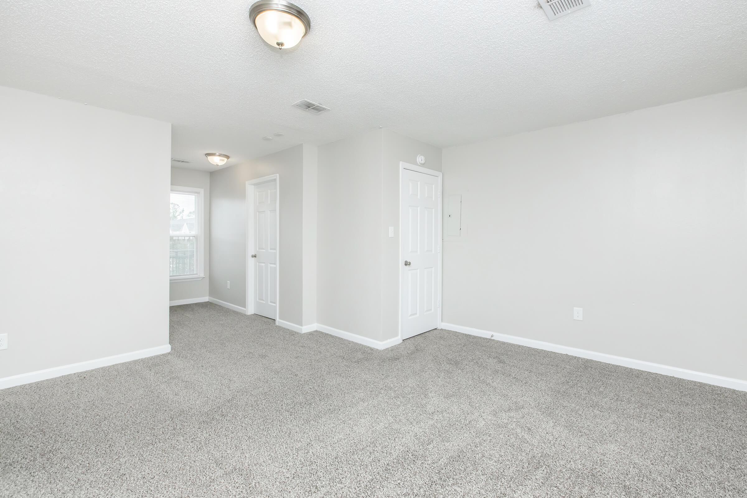 A spacious, newly carpeted room with light gray walls, featuring two light fixtures on the ceiling. There are two doorways, one leading to a bathroom and another to a windowed space, suggesting a bright and airy atmosphere. The floor is covered in plush beige carpeting.