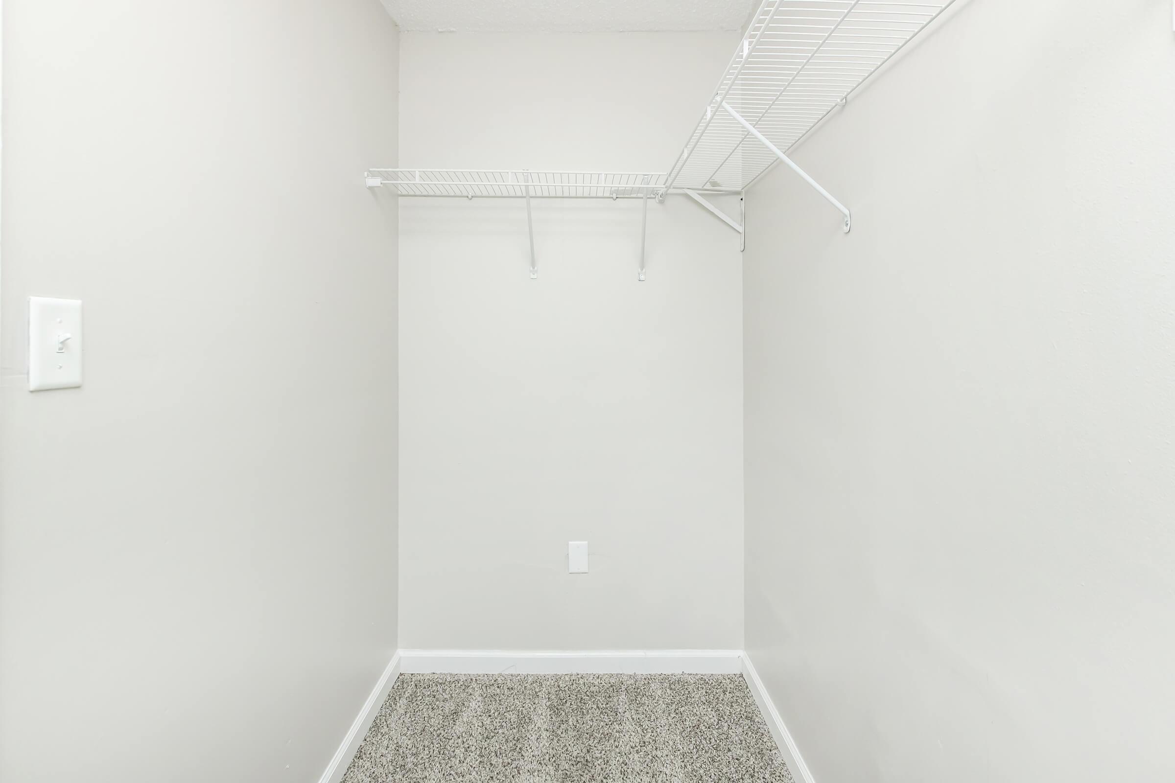 Empty closet with light gray walls and a carpeted floor. There is a wire shelving unit mounted on one side and a small electrical outlet on the wall. The space is well-lit and has no visible items or furniture, giving it a minimalist appearance.
