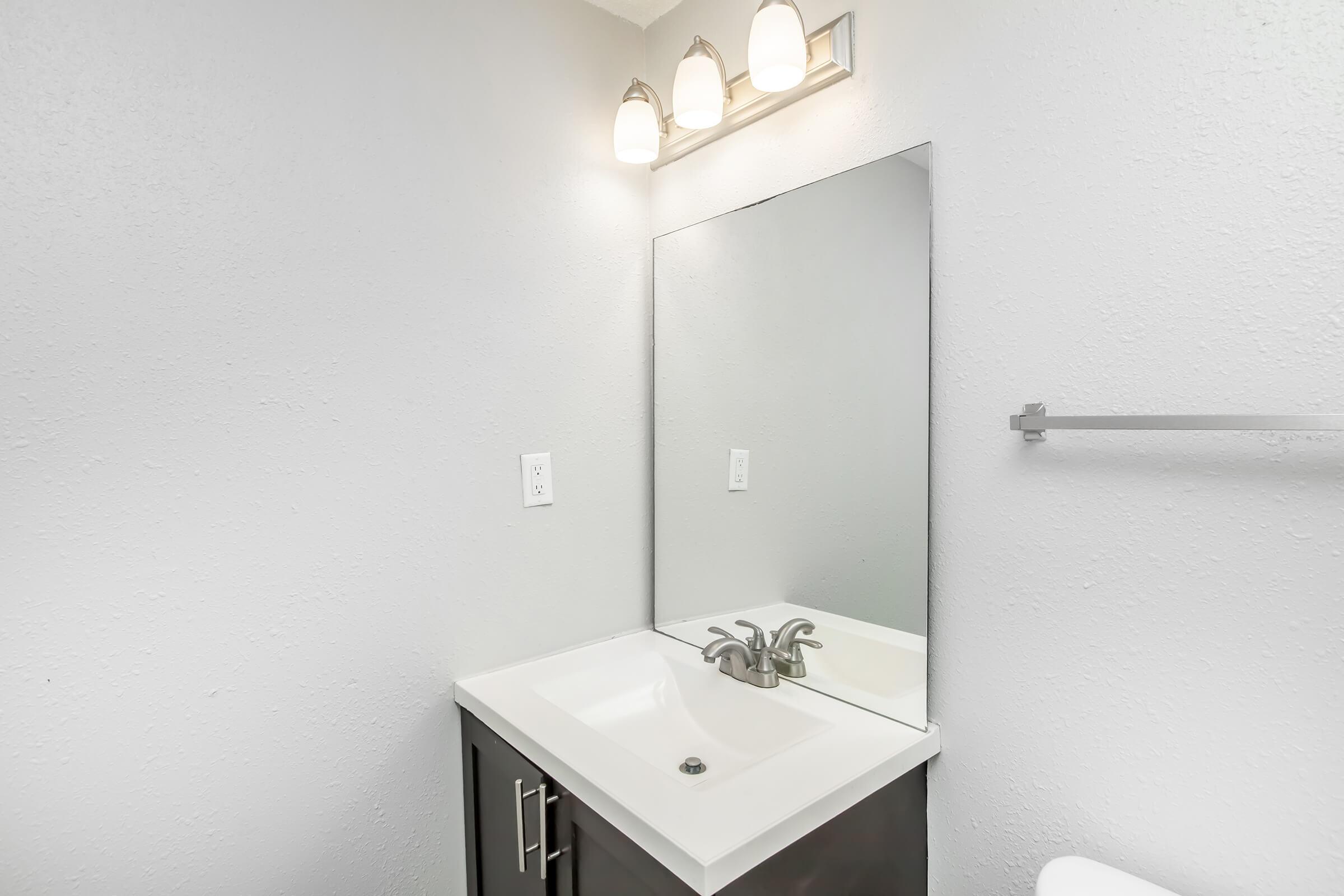 A clean and modern bathroom with a white vanity featuring a sink and a mirror above it. The space has three light fixtures above the mirror and a towel bar on the side wall. The walls are painted in a light color, enhancing the bright and airy feel of the room.