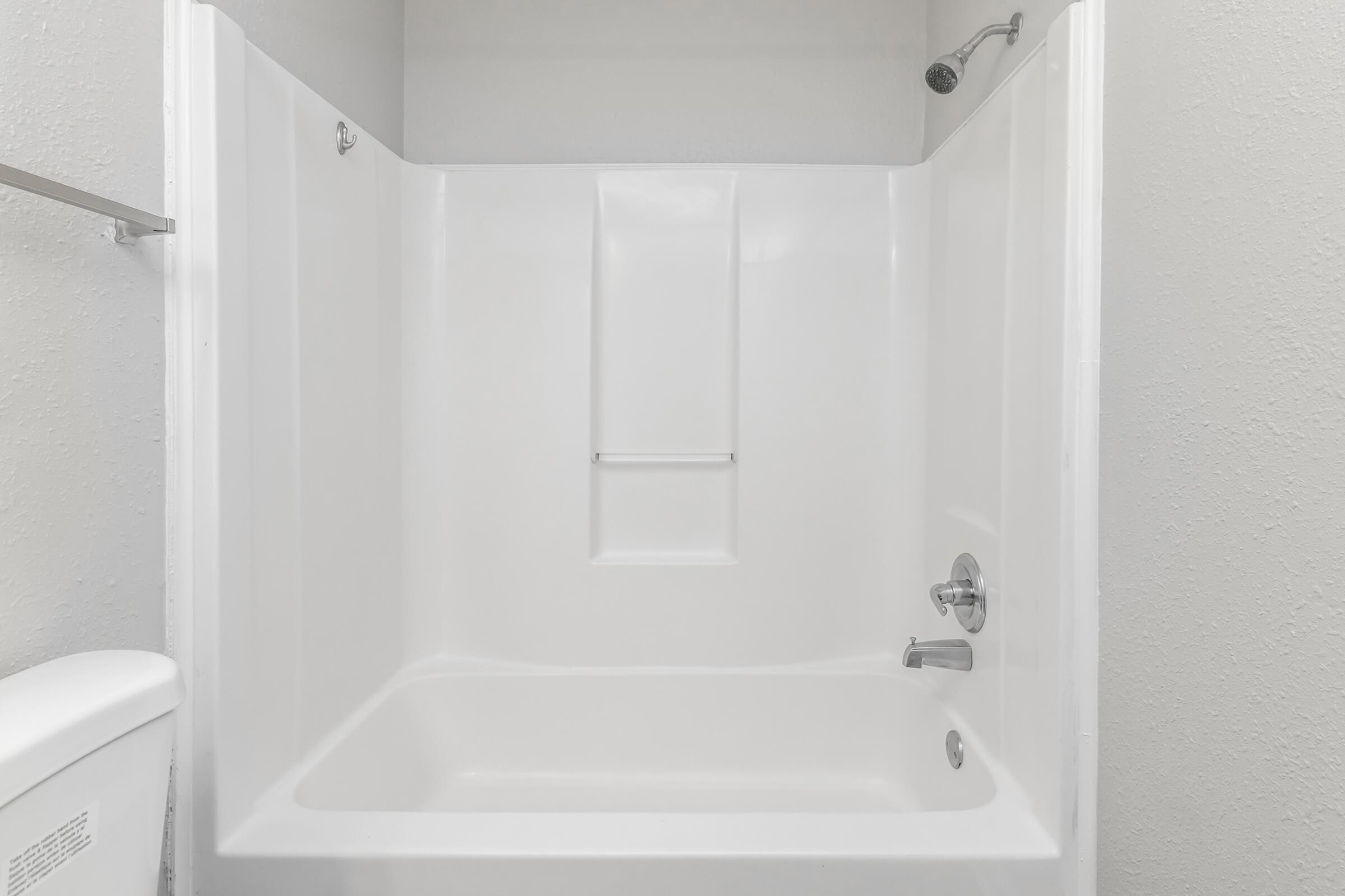 Clean and empty shower stall with a white acrylic surround, equipped with a faucet and hand-held showerhead. The background features a light-colored wall and a portion of a toilet in one corner of the frame. The overall aesthetic is simple and modern, suitable for a bathroom setting.