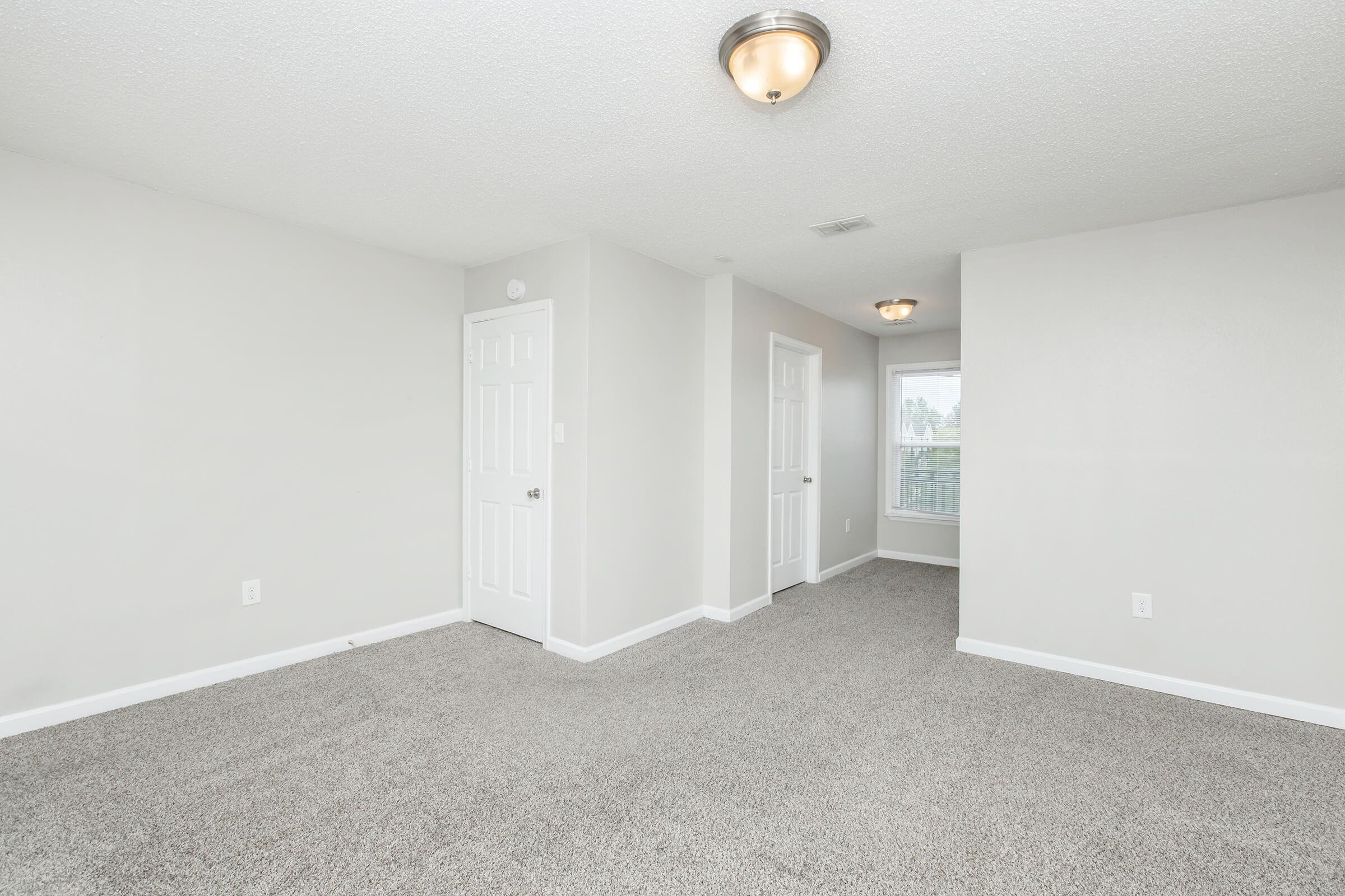 A spacious, well-lit room with light gray walls and carpeted flooring. Two white doors lead to adjacent spaces, and a window provides natural light from the right. The ceiling features modern light fixtures, contributing to a clean and inviting atmosphere.