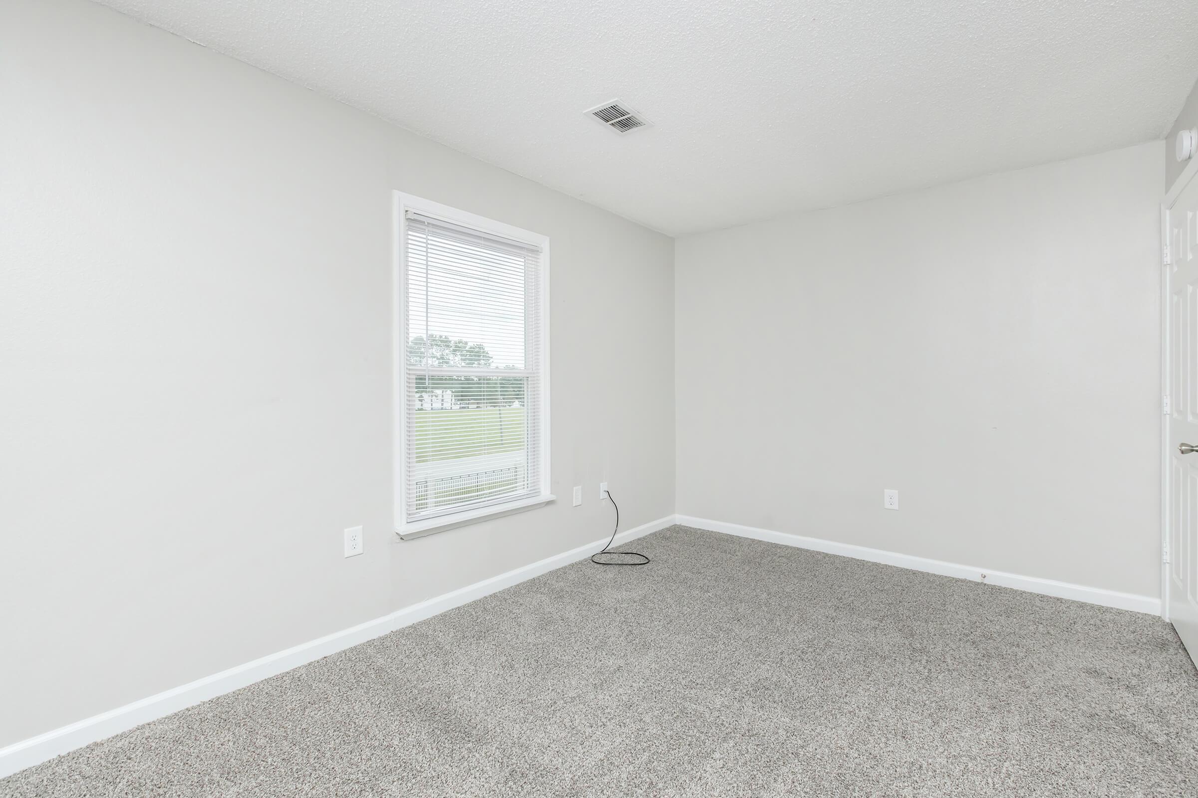 A small, empty room with light gray walls and plush beige carpeting. The room features a single window with white blinds, allowing natural light to enter. There's a visible electrical outlet near the floor, and the space appears clean and ready for furnishings.
