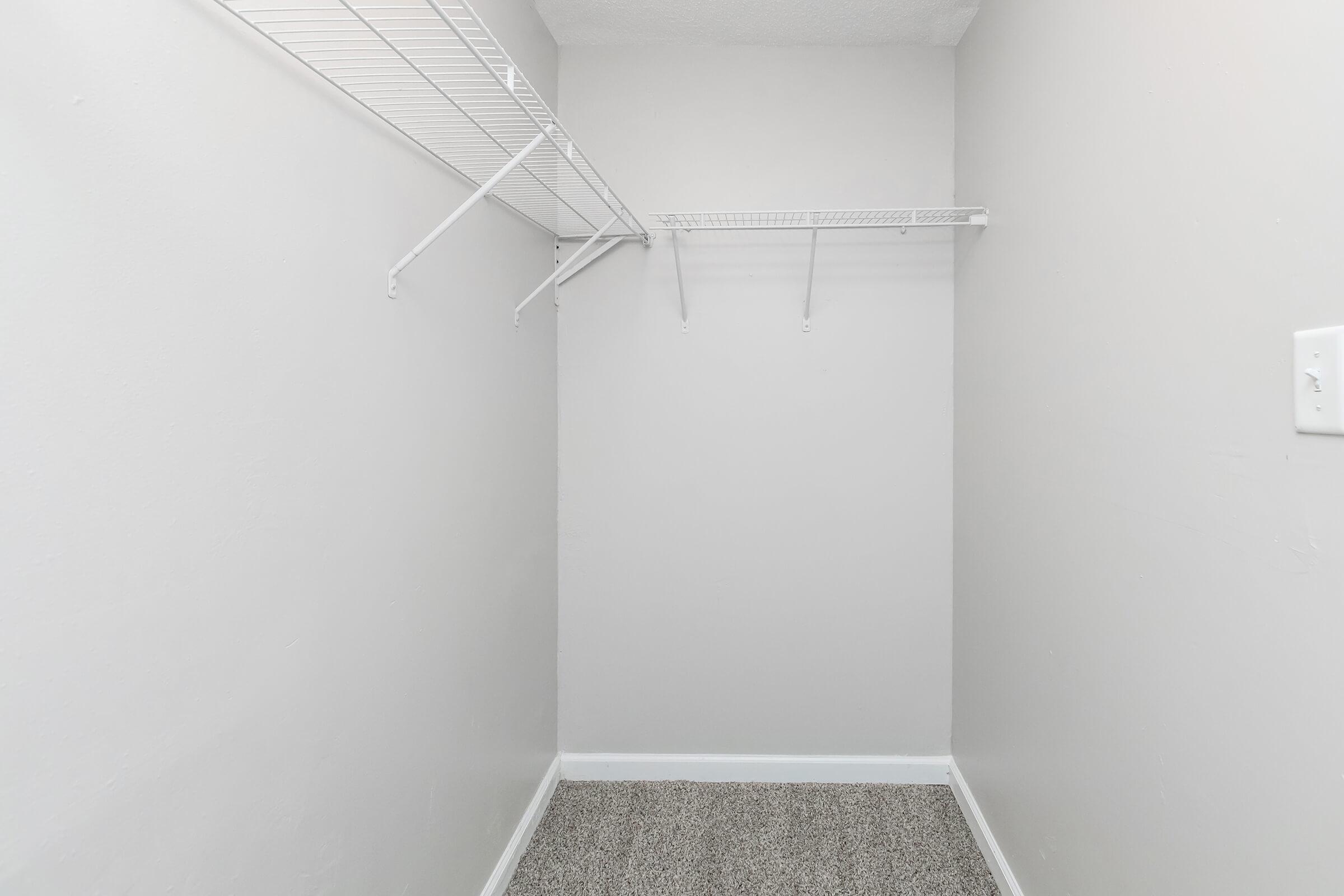 A small, empty closet with light gray walls and carpeted flooring. One side features a metal wire shelving unit mounted to the wall, while the opposite side has an additional shelf. The space appears clean and minimalistic, with no items stored inside.