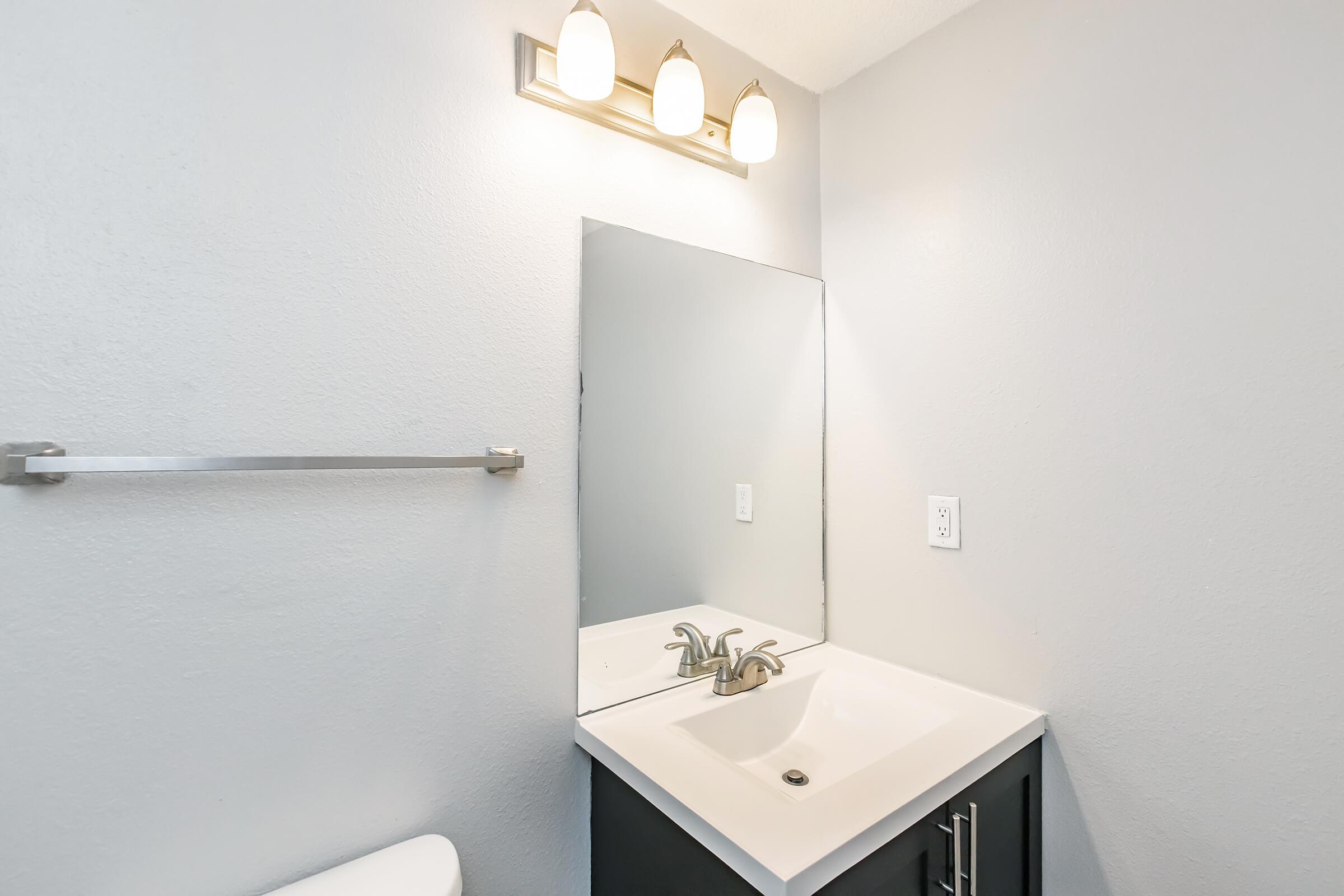A modern bathroom with a gray wall, featuring a white countertop sink with a chrome faucet, a wall-mounted mirror, and three light fixtures above. There is a towel bar made of metal, and a white toilet visible in the background. The overall design is clean and simple.
