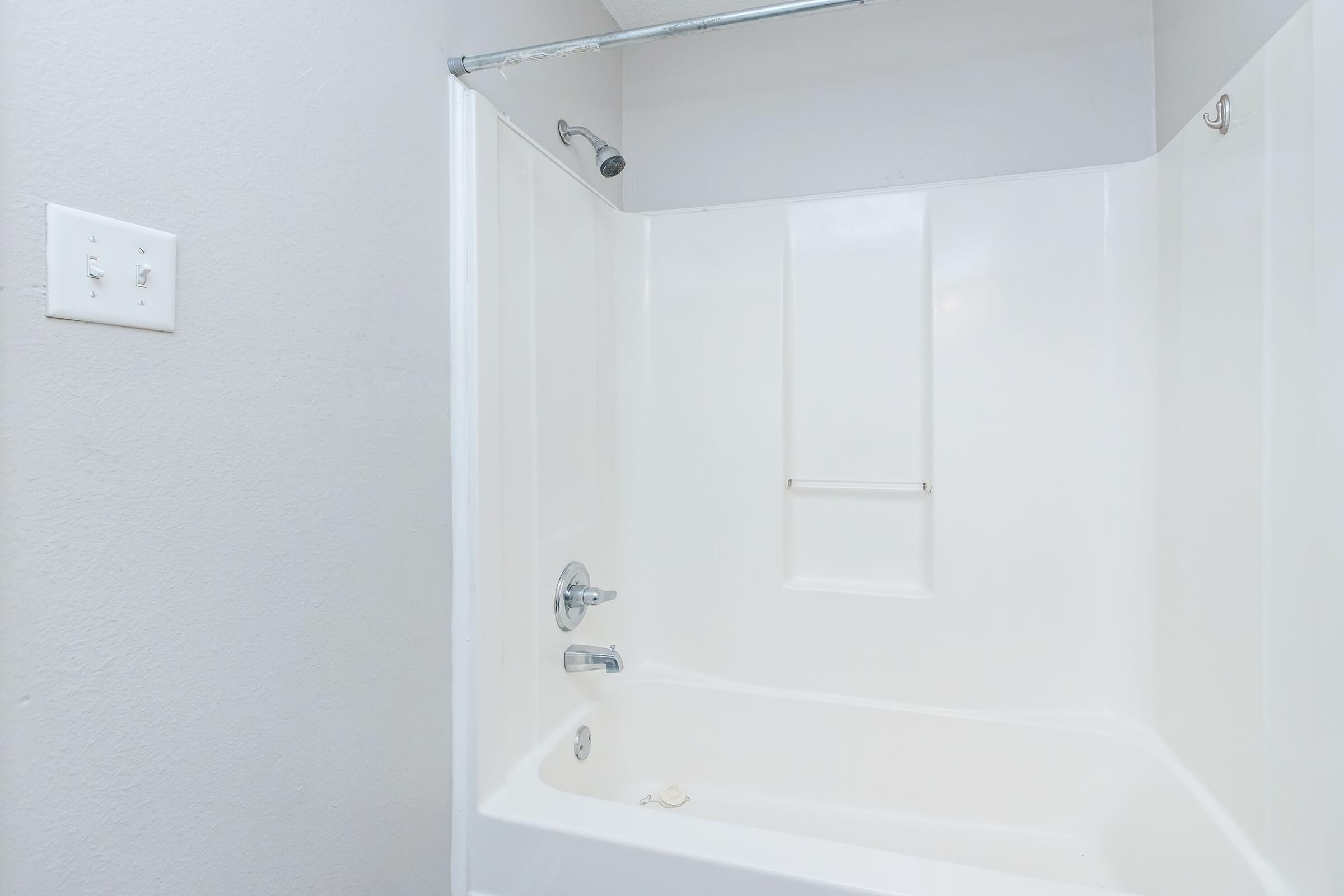 A clean, white bathtub and shower combo with a showerhead and a mounted towel bar. The walls are painted light gray, and there are electrical outlets on the left side. The overall look is simple and modern, emphasizing cleanliness and functionality.