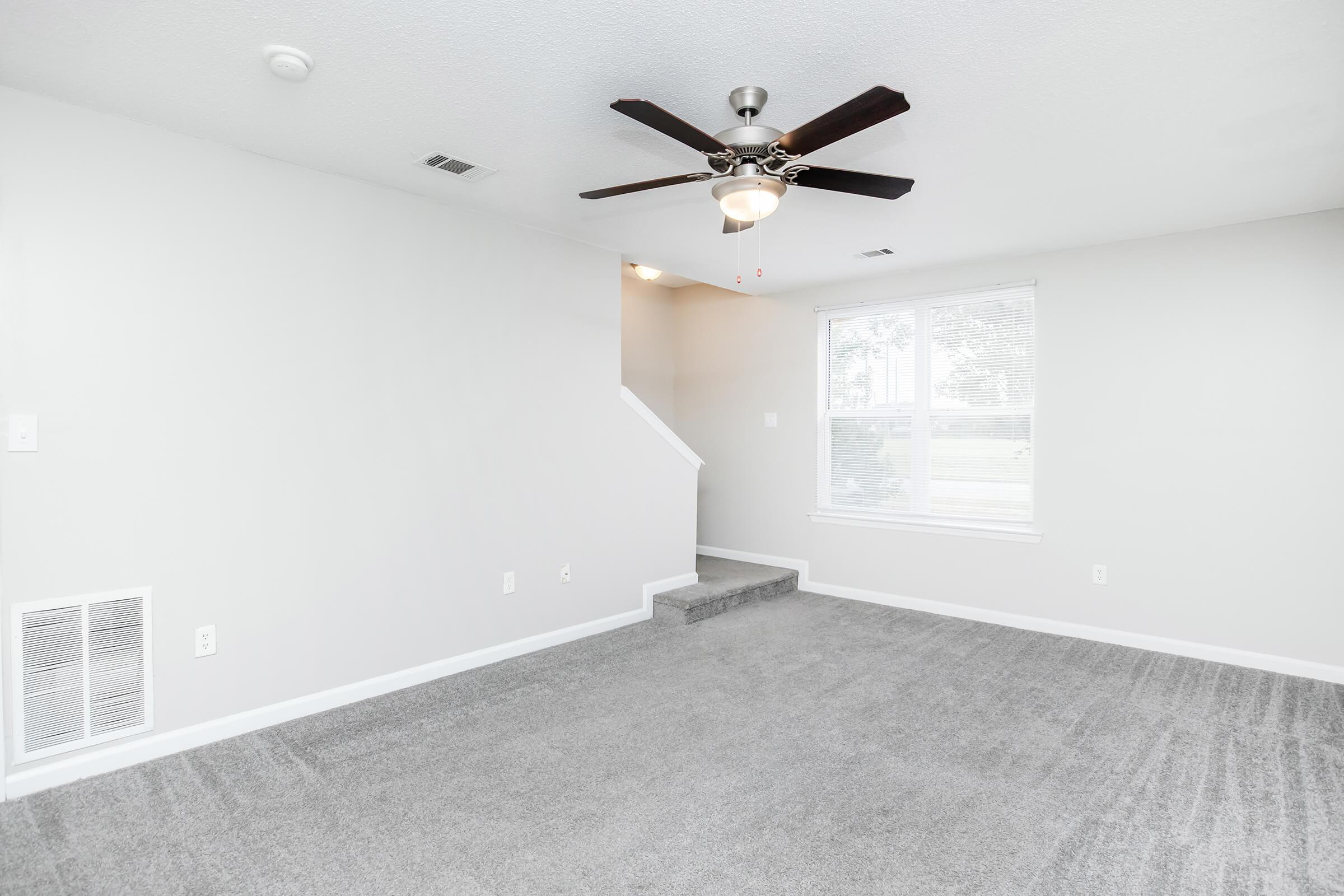 Empty living room with light gray carpet and painted walls. A ceiling fan is installed, and there are two windows allowing natural light. A staircase leads to another level in the background. The room is spacious and free of furniture, creating an open, inviting atmosphere.