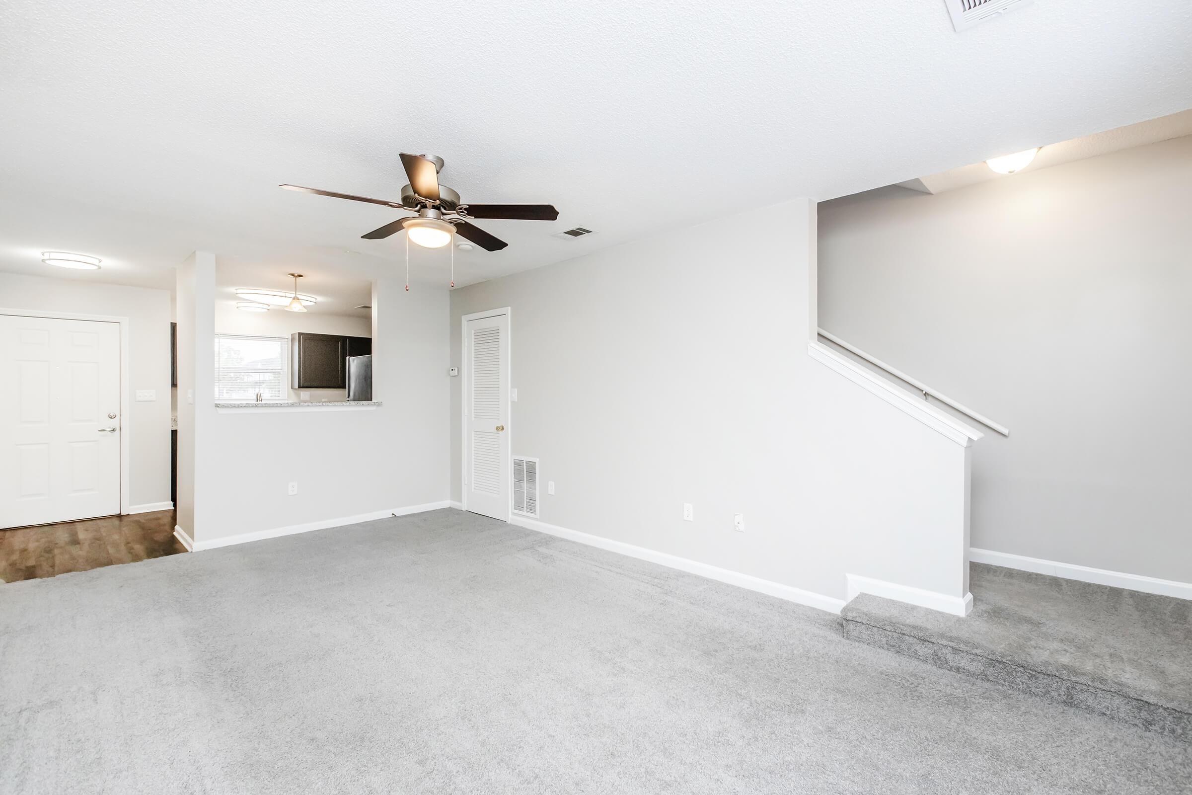 A spacious, empty living room with light gray carpet and walls. There is a ceiling fan, a staircase leading to another level, and an open view towards a kitchen area with dark cabinetry in the background. Natural light enters through the doorway, highlighting the room's neutral tones.