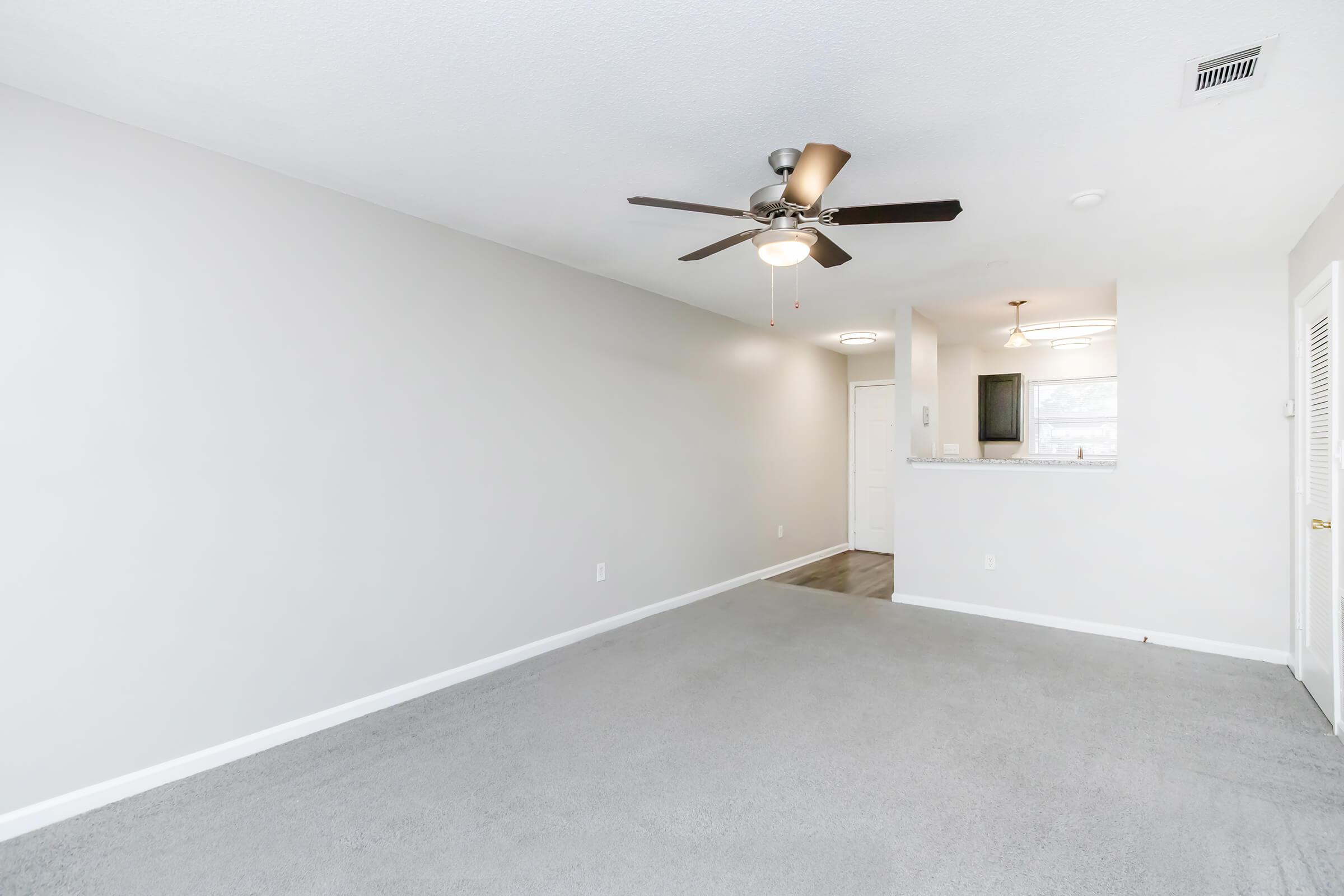 A spacious living room with light gray walls and carpet flooring. A ceiling fan is installed, and there is an open view towards a doorway and kitchen area with bar seating. Natural light enters the space, creating a bright and airy atmosphere.
