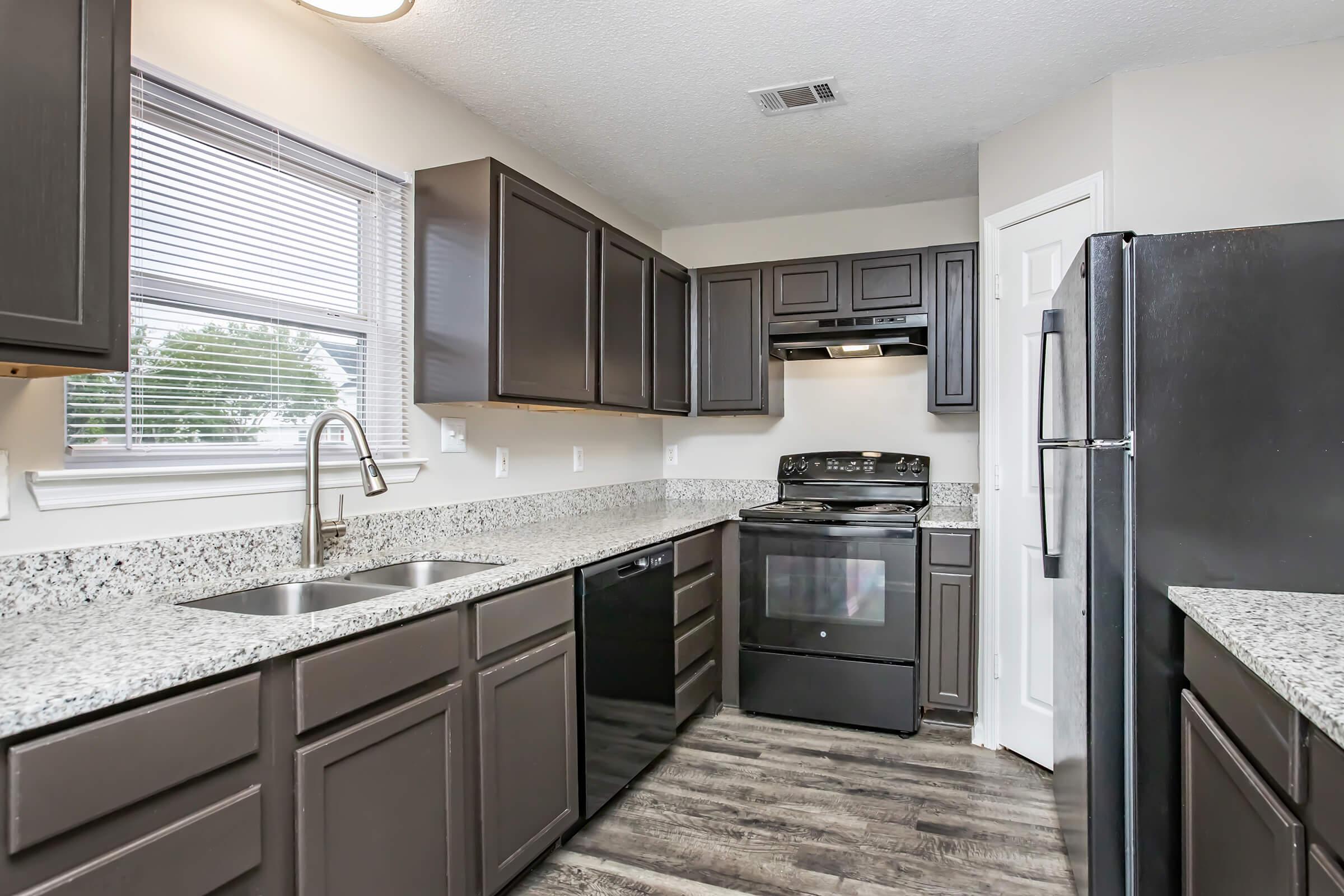 A modern kitchen featuring dark cabinetry and granite countertops. There is a stainless steel sink with a single faucet, a black refrigerator, an electric stove with an overhead microwave, and ample natural light coming through a window. The flooring is dark and complements the overall design.