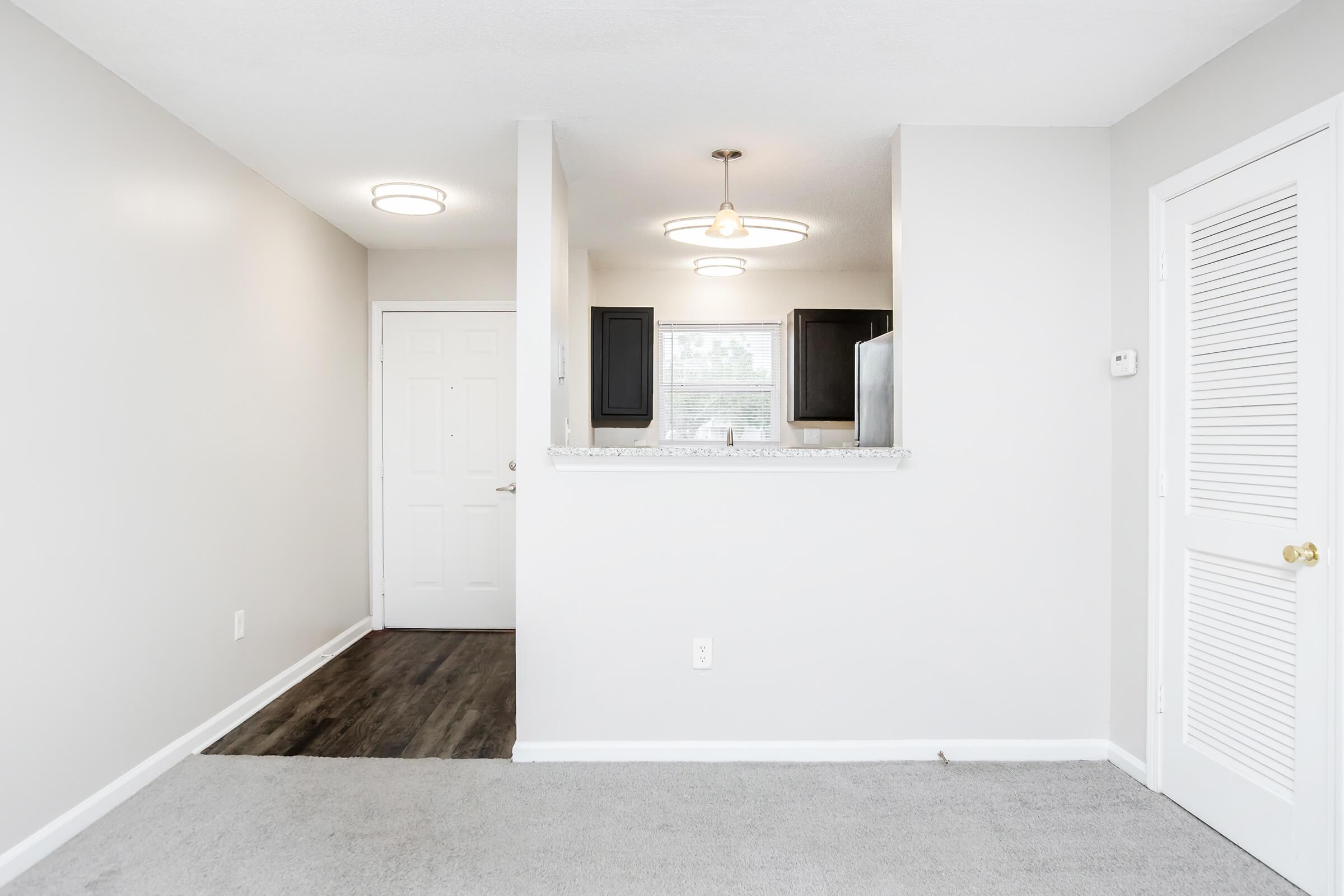 A bright and modern interior view of a room with light gray walls and carpet. To the left, there is a white door leading to an entrance. Ahead, a small kitchen area features dark cabinets and a window, with a refrigerator visible. Recessed lighting and a circular ceiling fixture provide illumination.