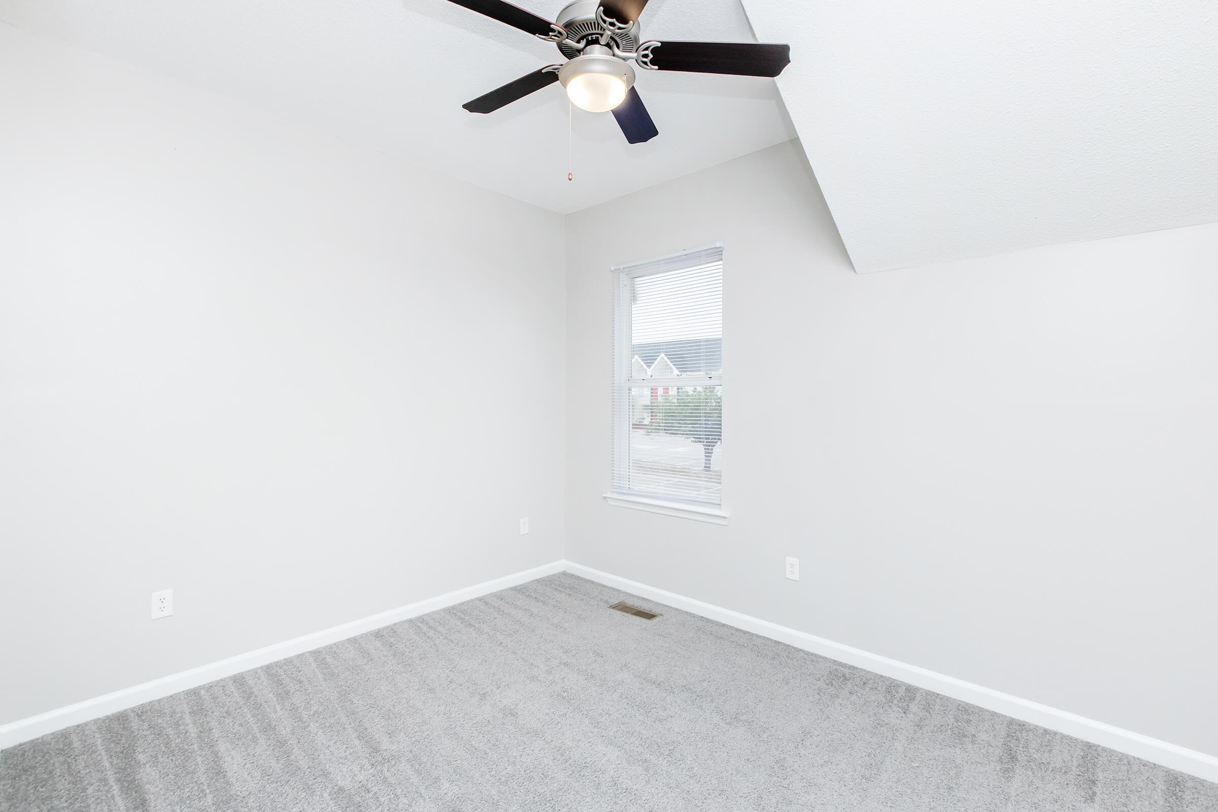 Empty room with light gray walls and soft gray carpet. Features a ceiling fan with a light fixture. A small window is partially open, allowing natural light to enter. The room has an angled ceiling, adding architectural interest.