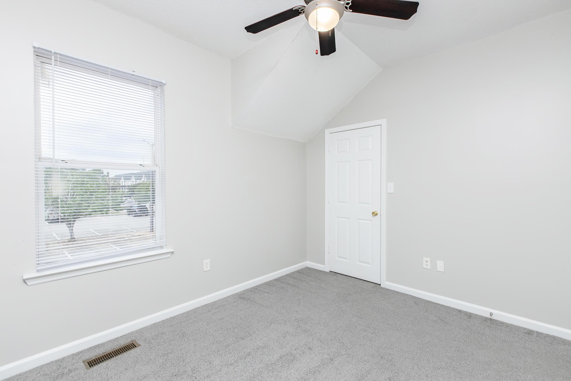 A vacant room with light gray walls and carpet. There’s a window with white blinds allowing natural light, and a ceiling fan. A white door is visible, and the overall space appears fresh and well-maintained. The sloped ceiling adds character to the room.