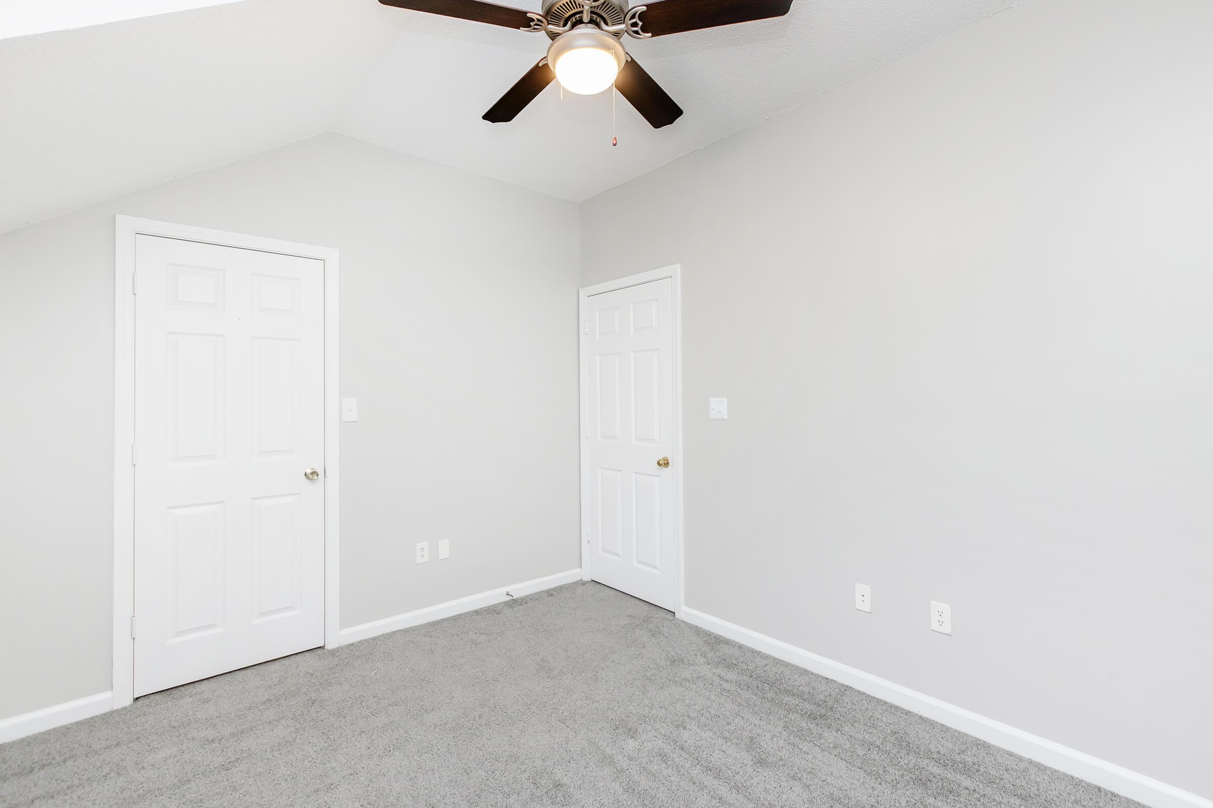 A simple interior room featuring light gray walls and fresh carpeting. There is a ceiling fan with a light fixture, a single window, and two closed white doors on the left and right walls. The space is bright and uncluttered, making it suitable for various uses.