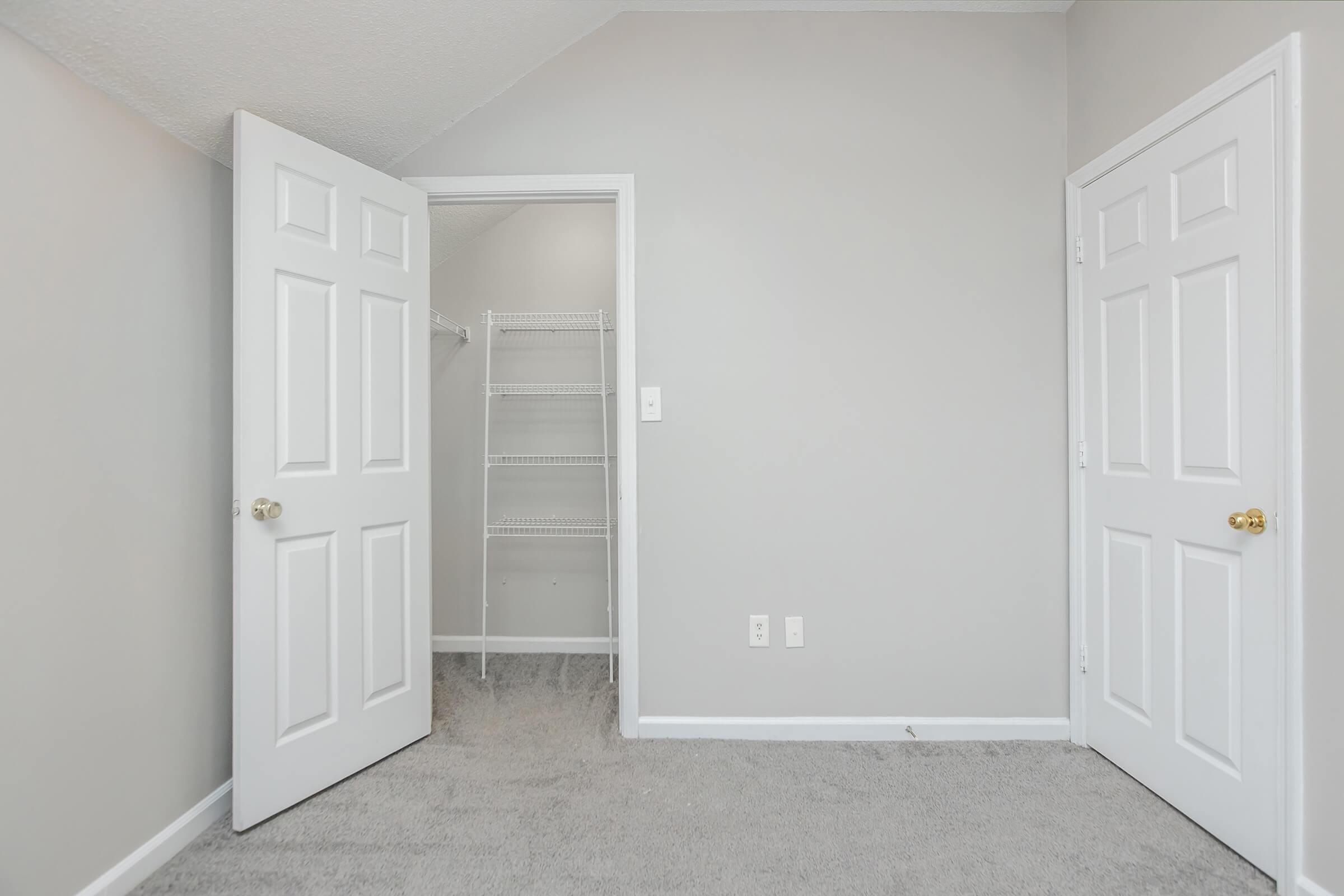 A simple, empty room with light gray walls and carpeting. Two white doors are slightly open: one leads to a small closet with a wire shelf, and the other is a standard interior door. The ceiling is slanted, and the overall atmosphere is bright and minimalist.