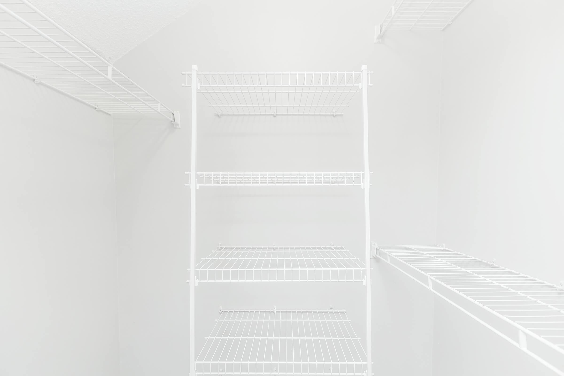 Empty white shelves in a minimalistic storage space. The shelves are made of wire and are mounted on white walls, creating a clean and spacious appearance. The background is bright and uncluttered, emphasizing the simplicity of the shelving design.