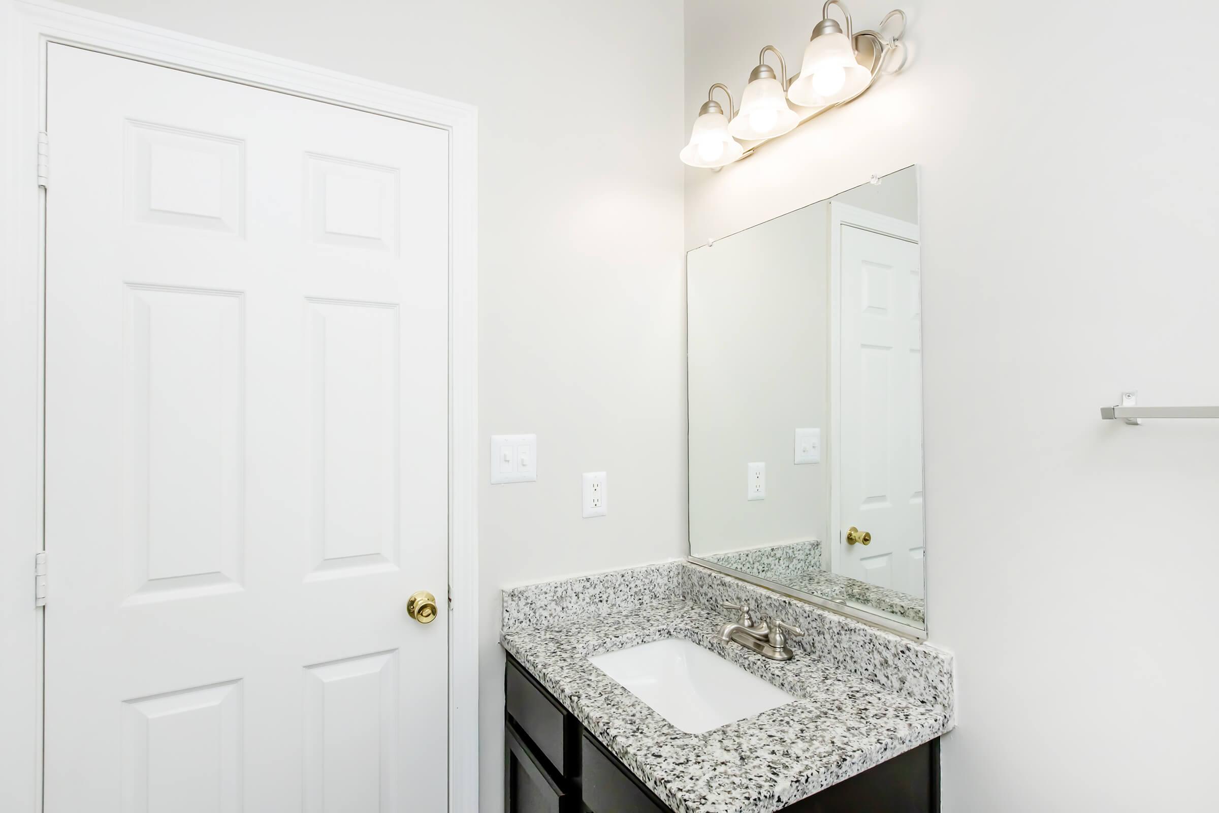 A clean bathroom featuring a sink with a granite countertop, a wall-mounted mirror, and a light fixture above. The door is white and closed, and a towel bar is visible on the wall. The overall aesthetic is bright and modern.
