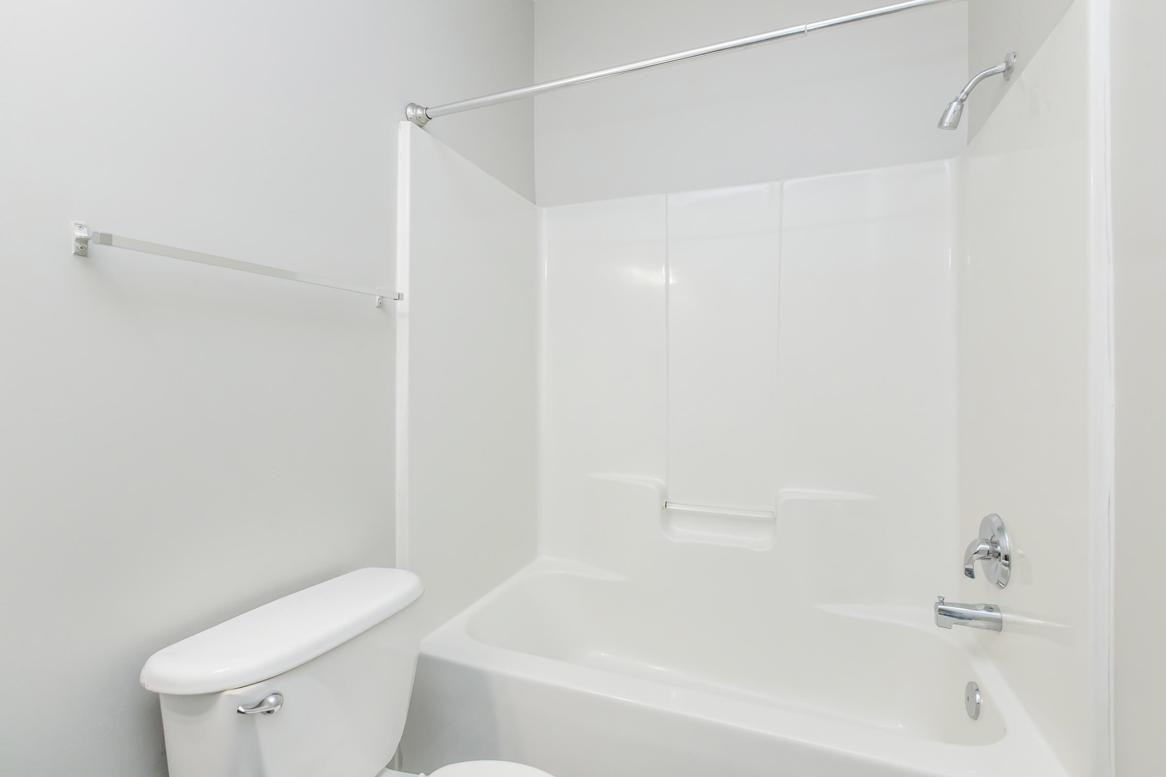 A clean, modern bathroom featuring a white bathtub with a showerhead above, a toilet beside it, and a simple chrome towel bar on the wall. The walls are painted a light gray, creating a bright and spacious atmosphere.