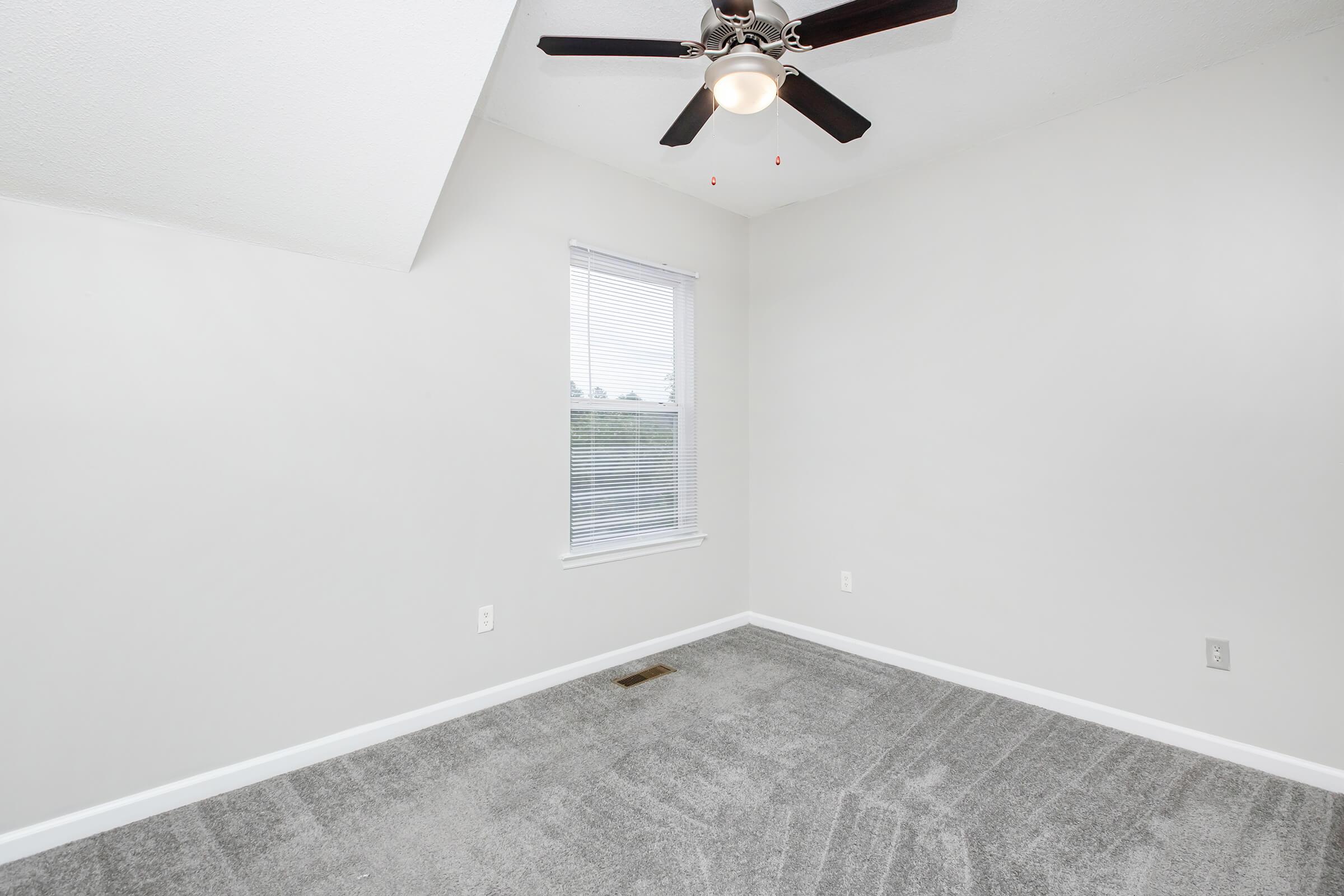A small, empty room with light gray walls and plush gray carpet. It features a ceiling fan with lights, a single window with blinds, and an angled ceiling. The space is uncluttered and ready for furniture or decoration.