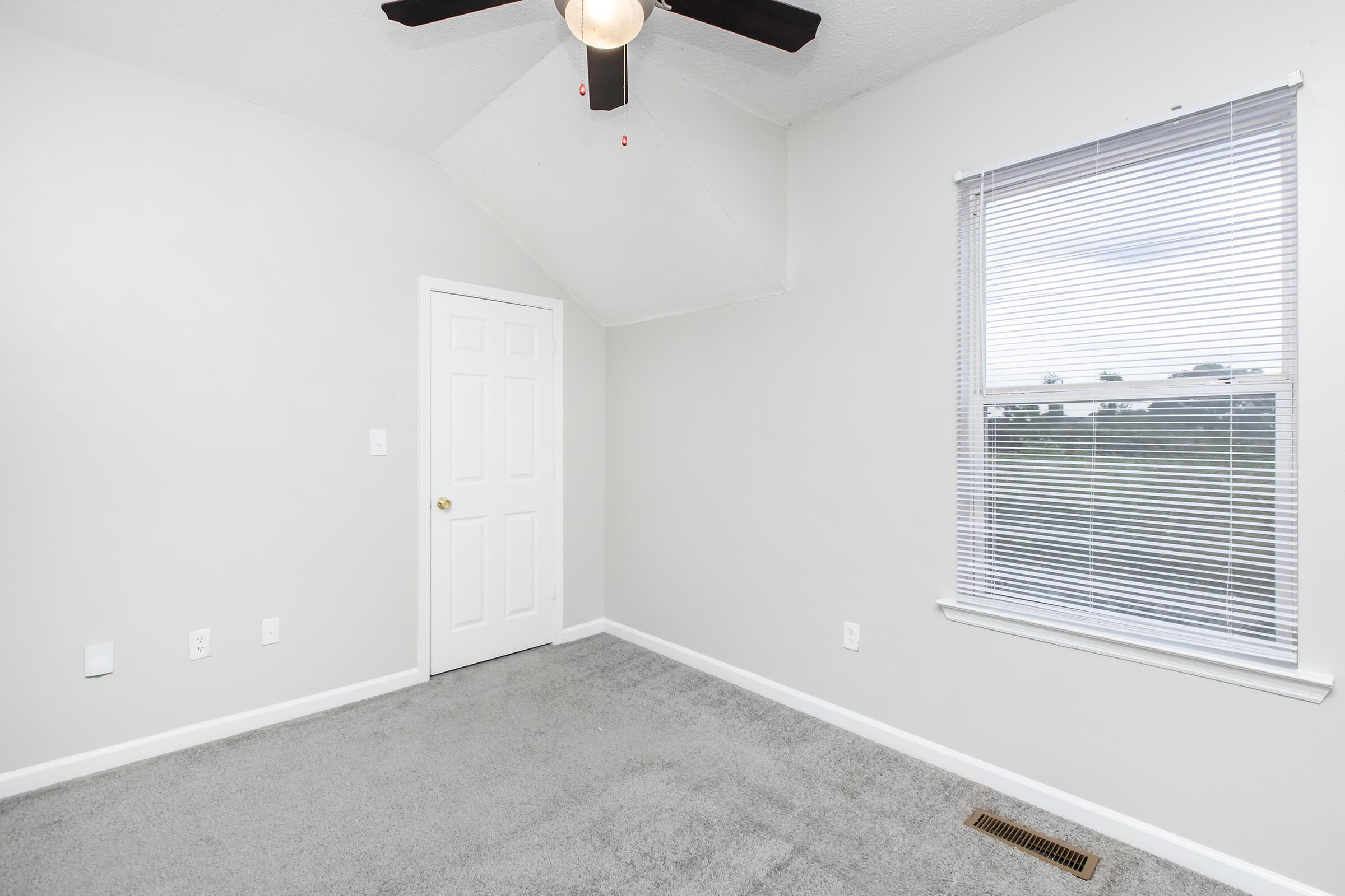 A light-colored room with gray carpet, featuring a ceiling fan and a window with blinds. There is a closed door on one wall and a small vent on the floor. The walls are painted in a light shade, creating a bright and spacious appearance.