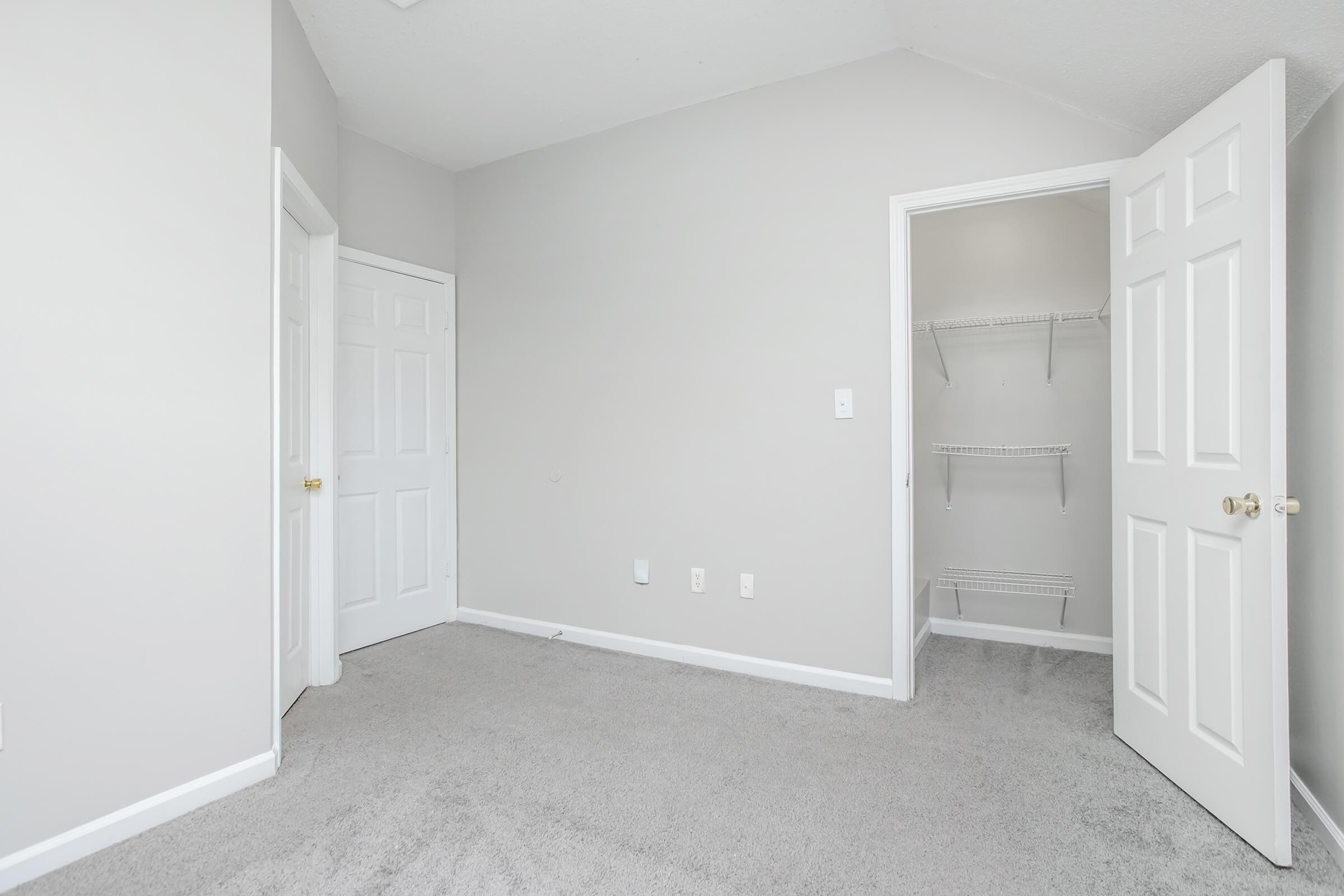 Empty room with gray walls and carpet. One door is ajar, revealing a small closet with wire shelving. The opposite wall features another closed door. The overall ambiance is clean and simple, with no furniture or decorations present.