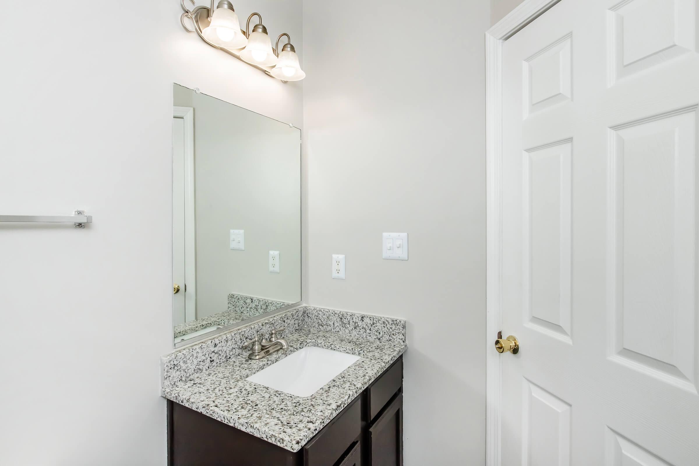 A clean bathroom with a granite countertop sink, a mirror above it, and a light fixture with three bulbs. The wall is painted in a light color, and a closed door is visible on the right side, showcasing a simple white design. A towel rack is mounted on the wall beside the sink.