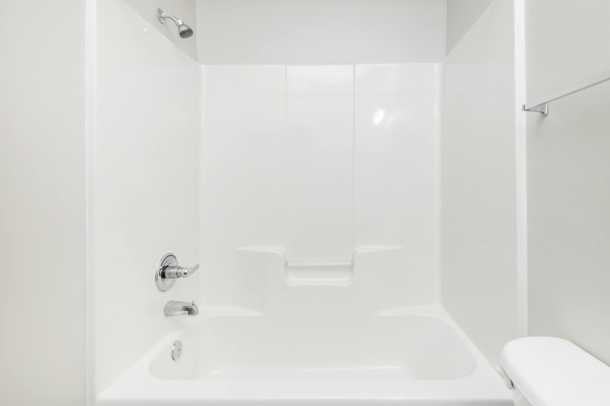 A clean, modern bathroom featuring a white fiberglass tub and shower unit. The walls are smooth and white, reflecting light, with a chrome showerhead and faucet. A small ledge is visible for toiletries, and a toilet is positioned to the right, creating a simple and functional space.