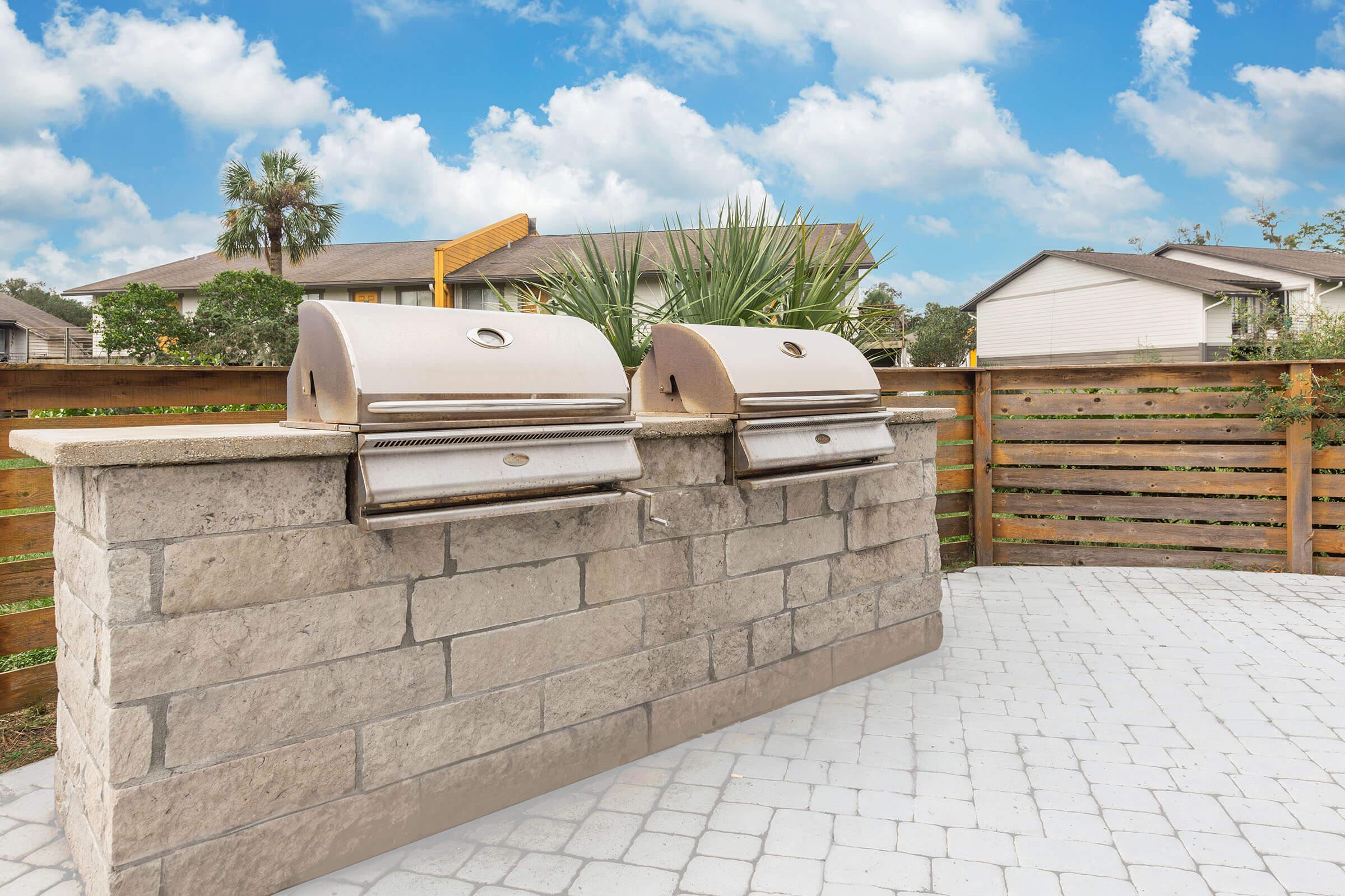 Two stainless steel grills built into a stone structure, set in a backyard with a paved surface. Behind the grills is a wooden fence and a small palm tree, against a backdrop of blue sky with fluffy white clouds and suburban houses.