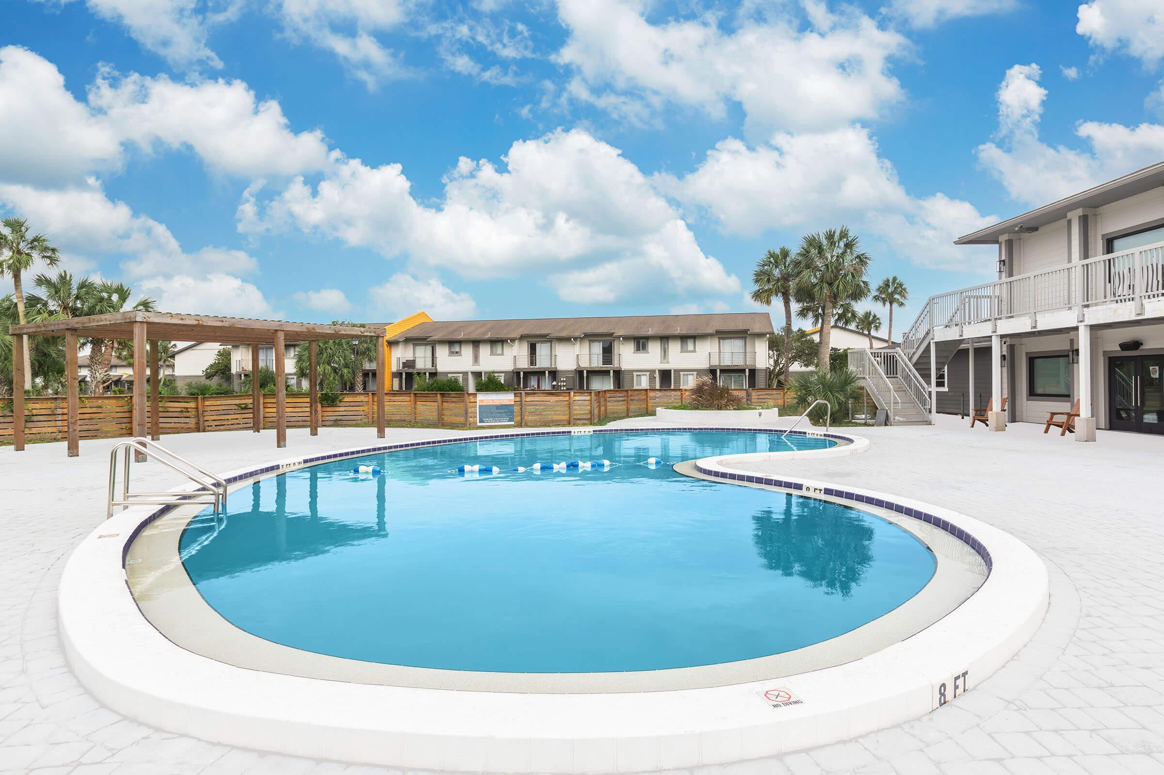 A clear blue swimming pool with a unique shape, surrounded by white deck tiles. Palm trees are visible in the background, along with a multi-story building that houses several balconies. The sky is partly cloudy, adding a bright atmosphere to the scene.