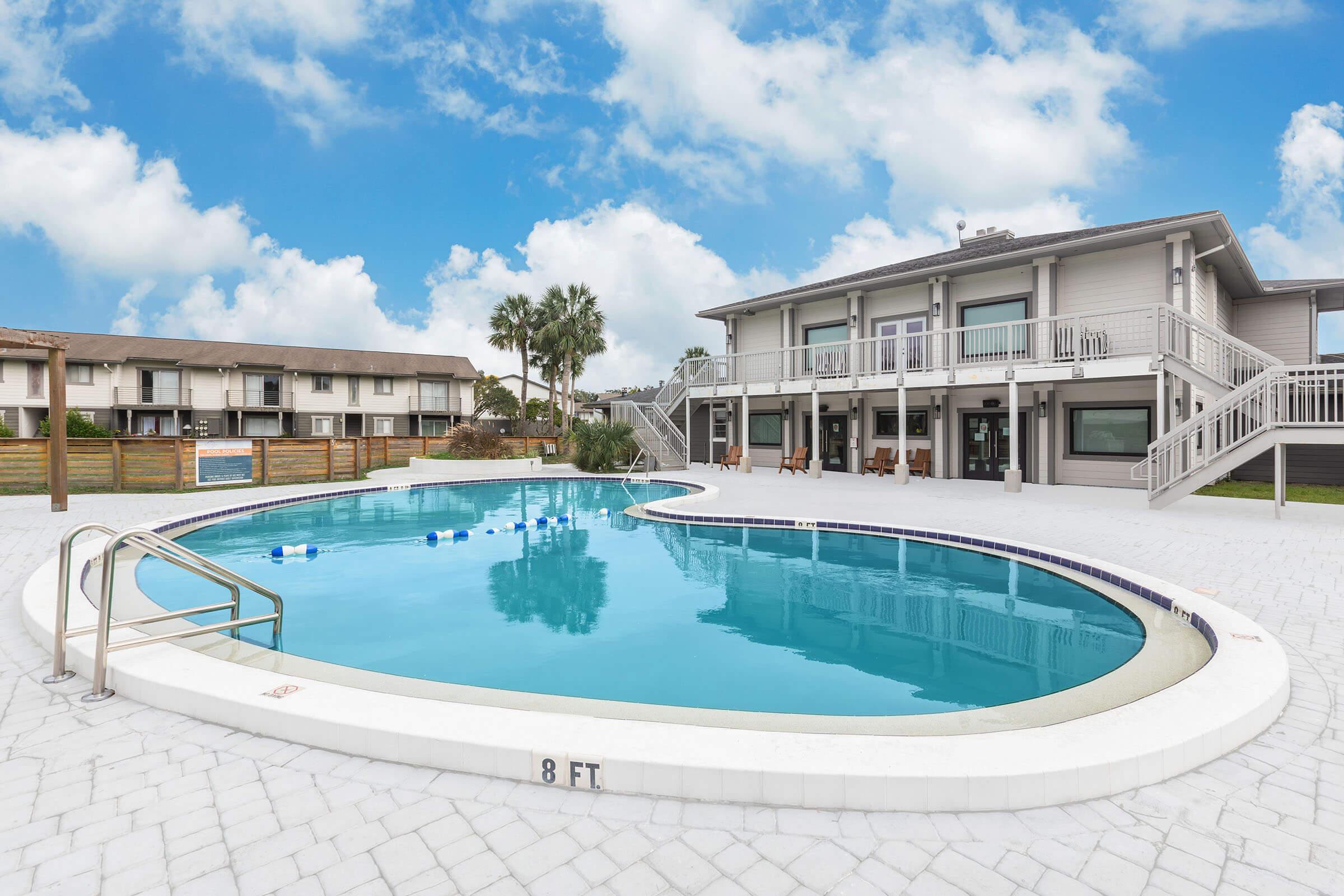 A large, inviting swimming pool with a depth of 8 feet, surrounded by a spacious deck featuring lounge chairs. In the background, a two-story building with multiple balconies and palm trees can be seen against a bright blue sky with fluffy white clouds.