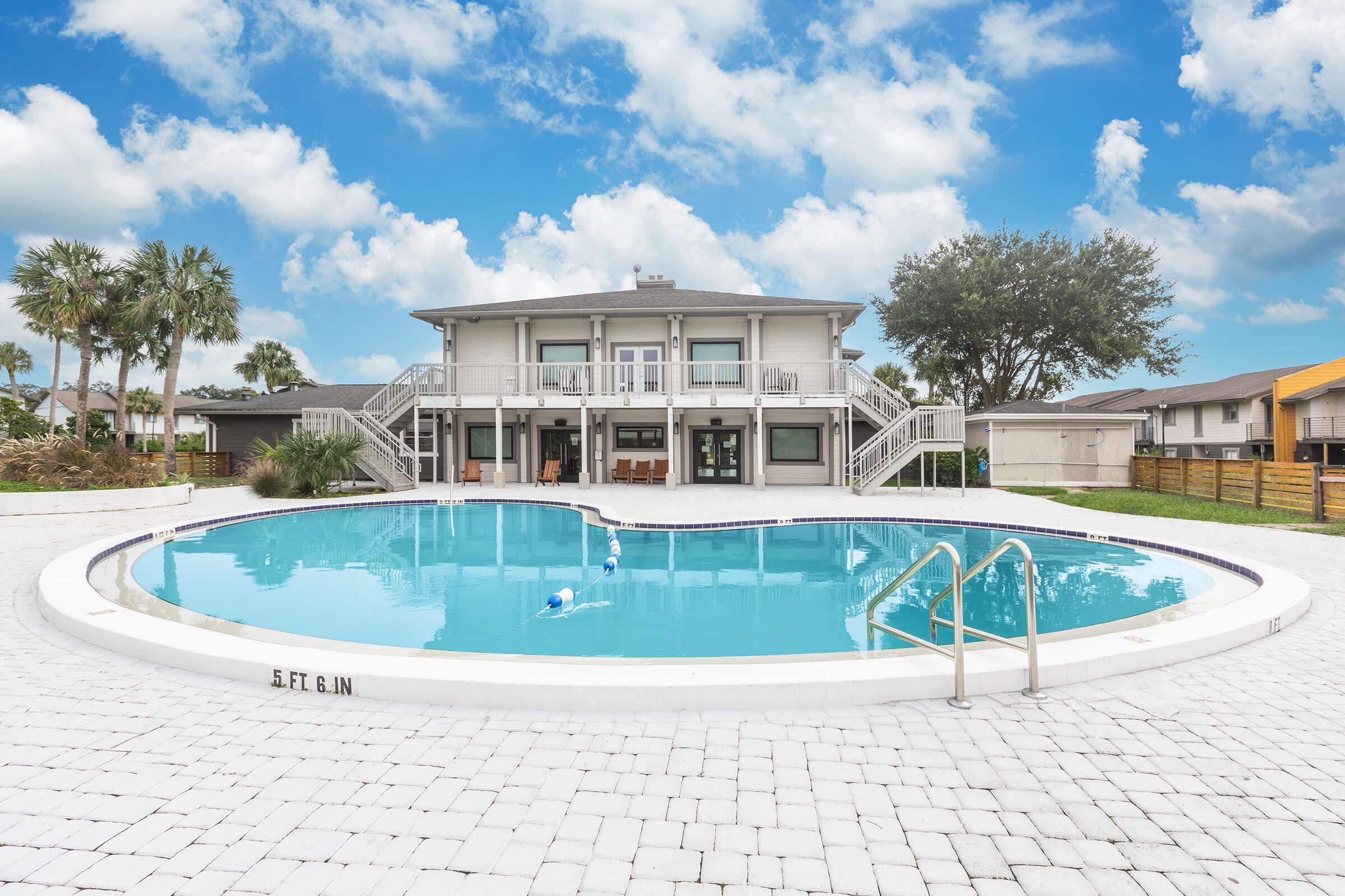 A heart-shaped swimming pool in front of a large, modern building with multiple balconies. The pool is surrounded by palm trees and a bright blue sky with fluffy white clouds. The area is well-maintained with a white tiled deck.