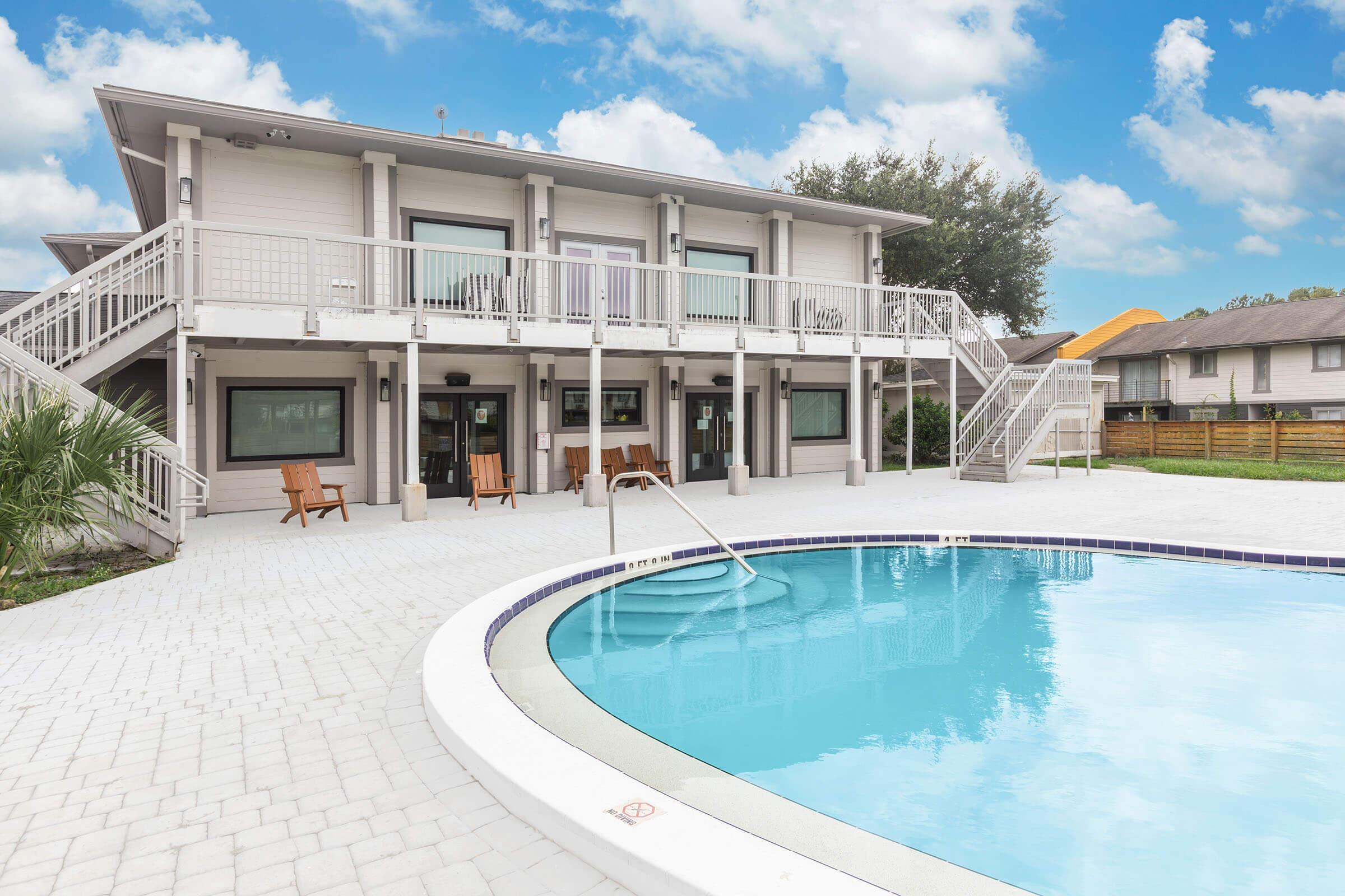 A modern two-story building with a spacious deck and multiple entrances overlooks a clear blue swimming pool. The surrounding area features palm trees and houses in the background under a partly cloudy sky. The pool area is paved with white tiles, creating a relaxing outdoor atmosphere.