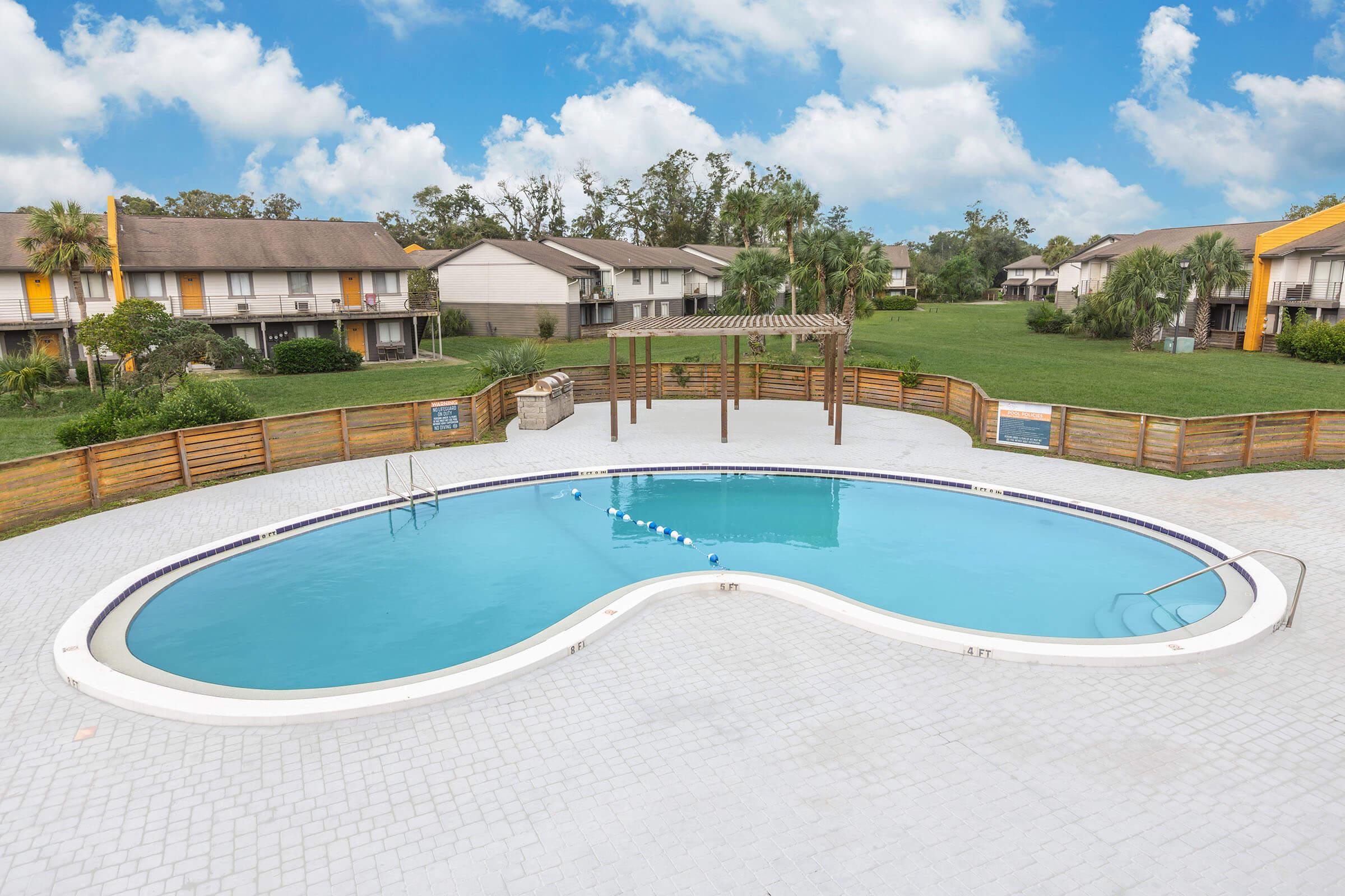 A clear blue swimming pool shaped like a figure eight, surrounded by a patio with light-colored pavers. There are lounge chairs nearby and a wooden pergola in the background. Residential buildings with brown roofs are visible in the distance, along with green grass and trees under a cloudy blue sky.