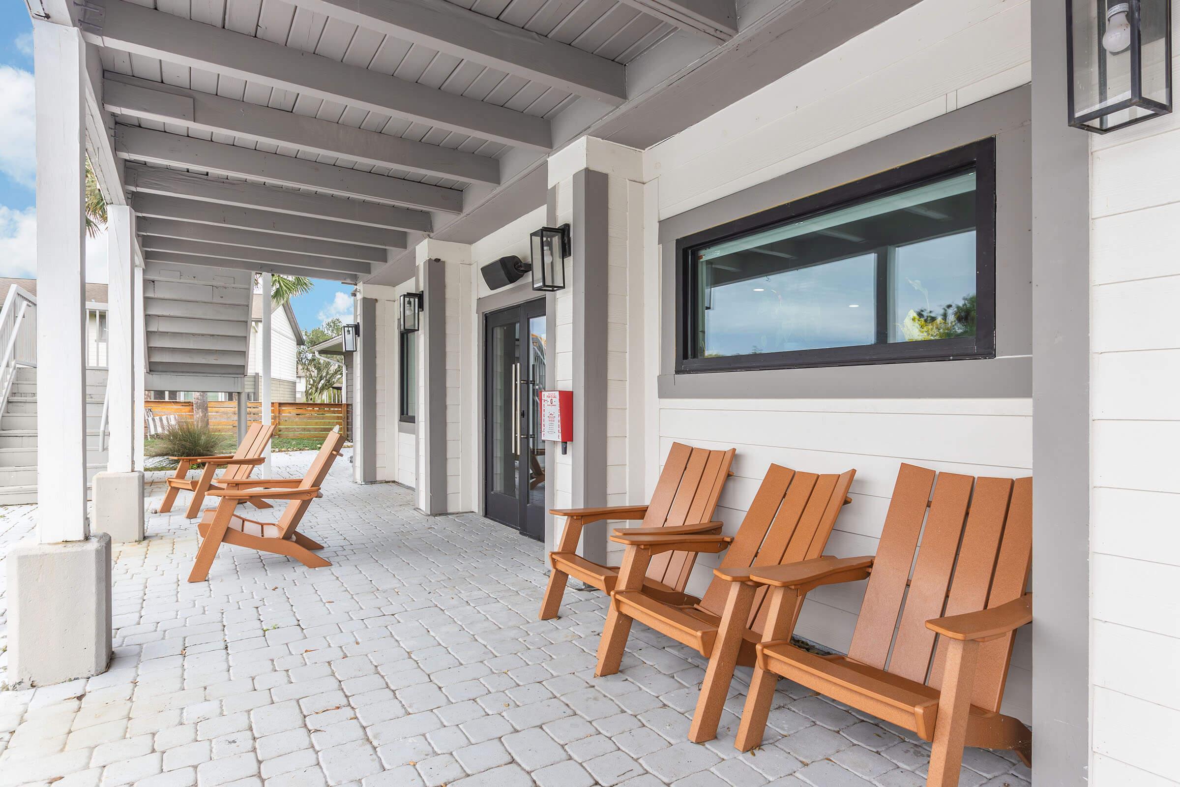 A view of a covered porch with light-colored wooden beams, featuring several wooden adirondack chairs arranged in a row on a stone-paved floor. The porch has a large window and modern lantern-style light fixtures, creating a welcoming outdoor space.