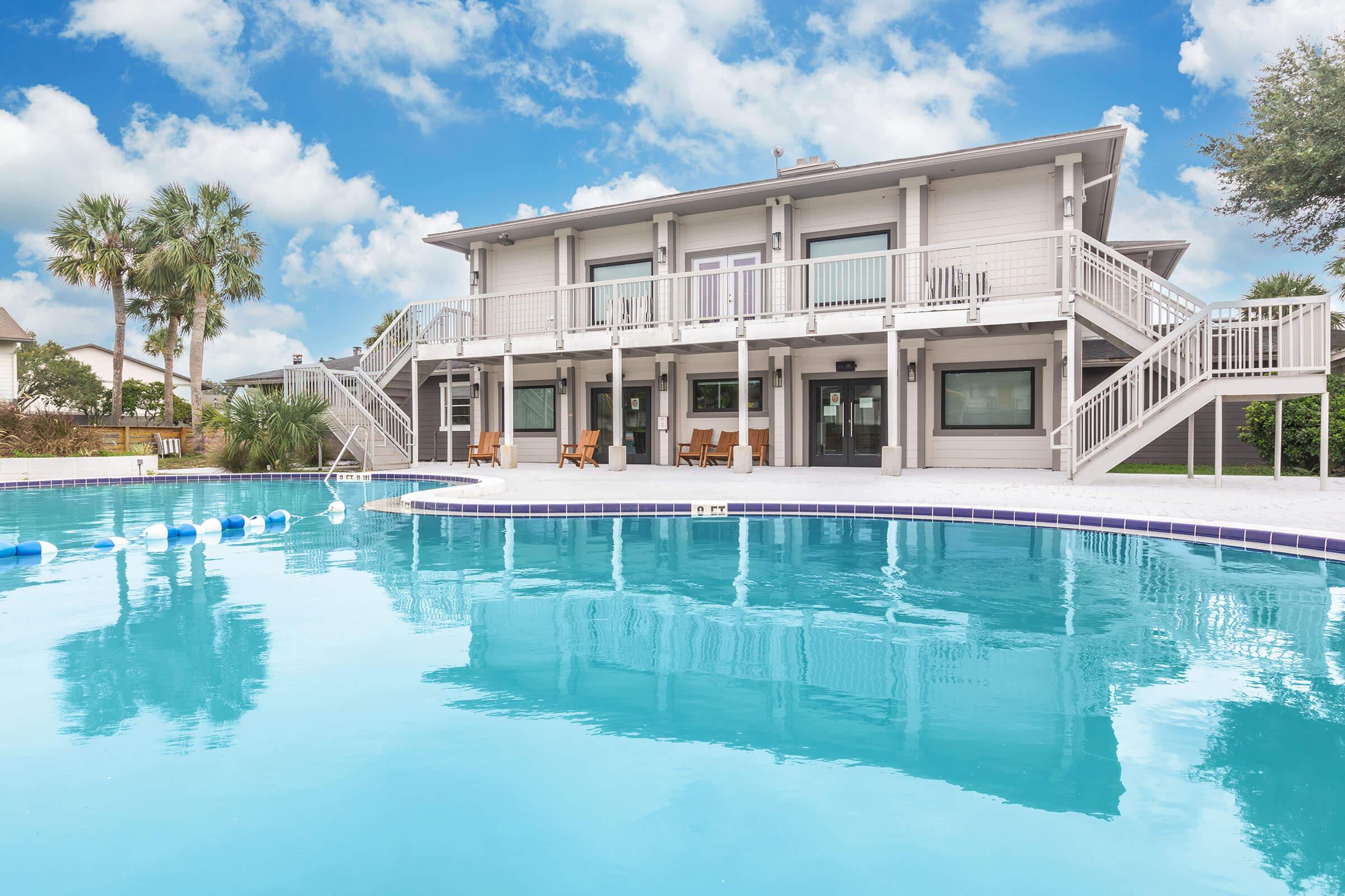 A spacious pool area featuring a crystal-clear blue pool in front of a two-story house with balconies. The house has large windows and a modern design, surrounded by palm trees and greenery. A few lounge chairs are positioned near the pool, and the sky is bright with scattered clouds.