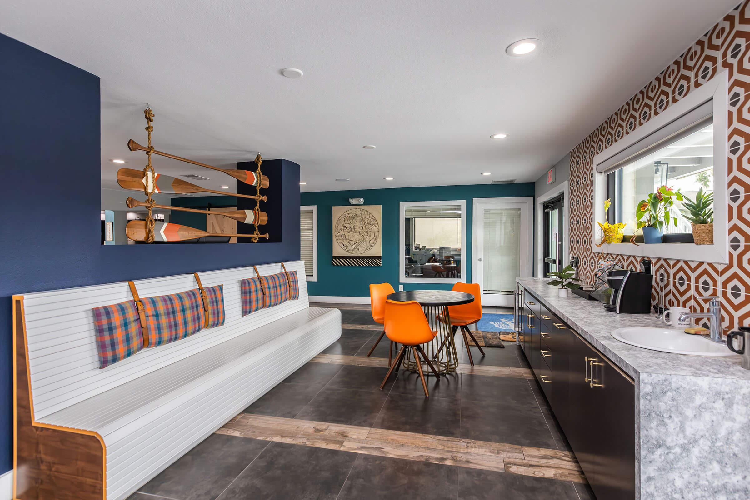 Interior view of a modern living space featuring a combination of navy blue walls and geometric patterned accents. There’s a cozy bench with plaid cushions, a round table with bright orange chairs, and a sleek kitchen area with dark cabinets. Sunlight streams in through large windows, enhancing the inviting atmosphere.