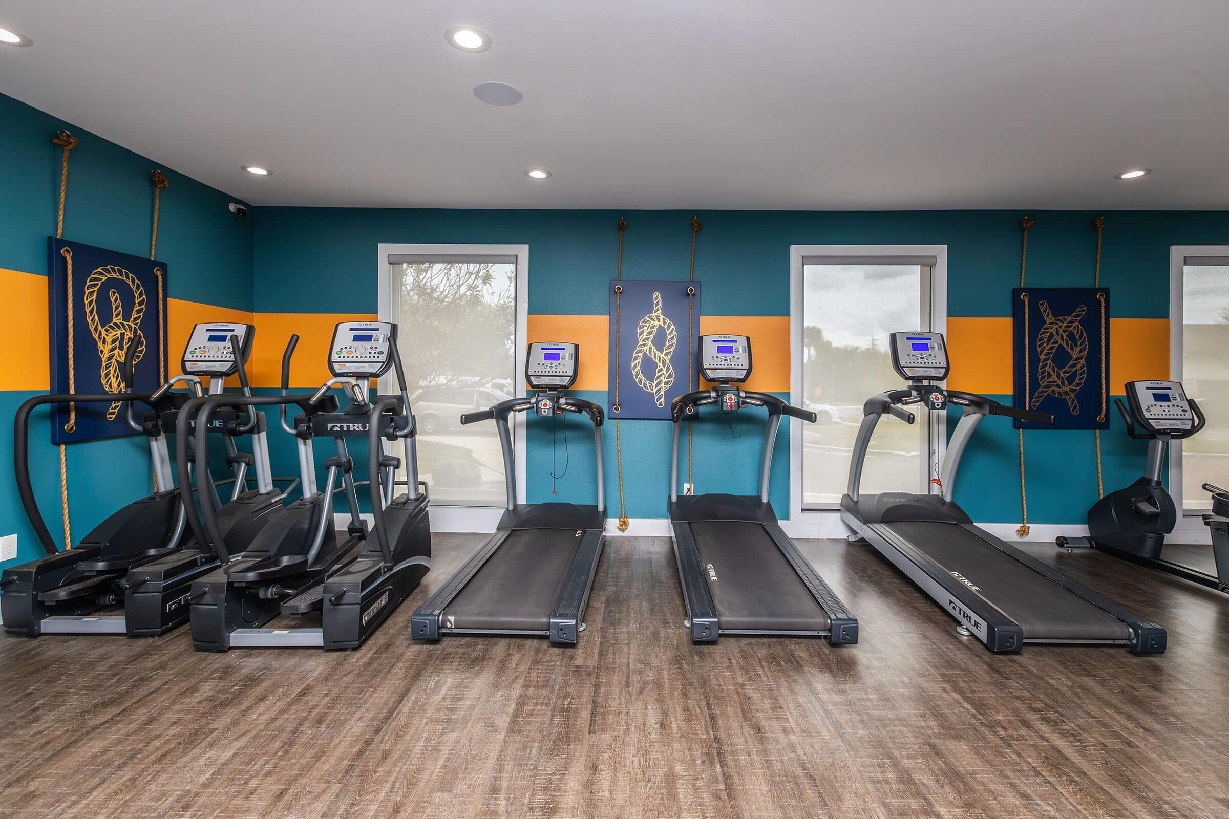 A fitness room featuring multiple treadmills and elliptical machines against a teal and orange wall. Brightly lit with windows, the space is equipped for exercise, promoting a healthy lifestyle.