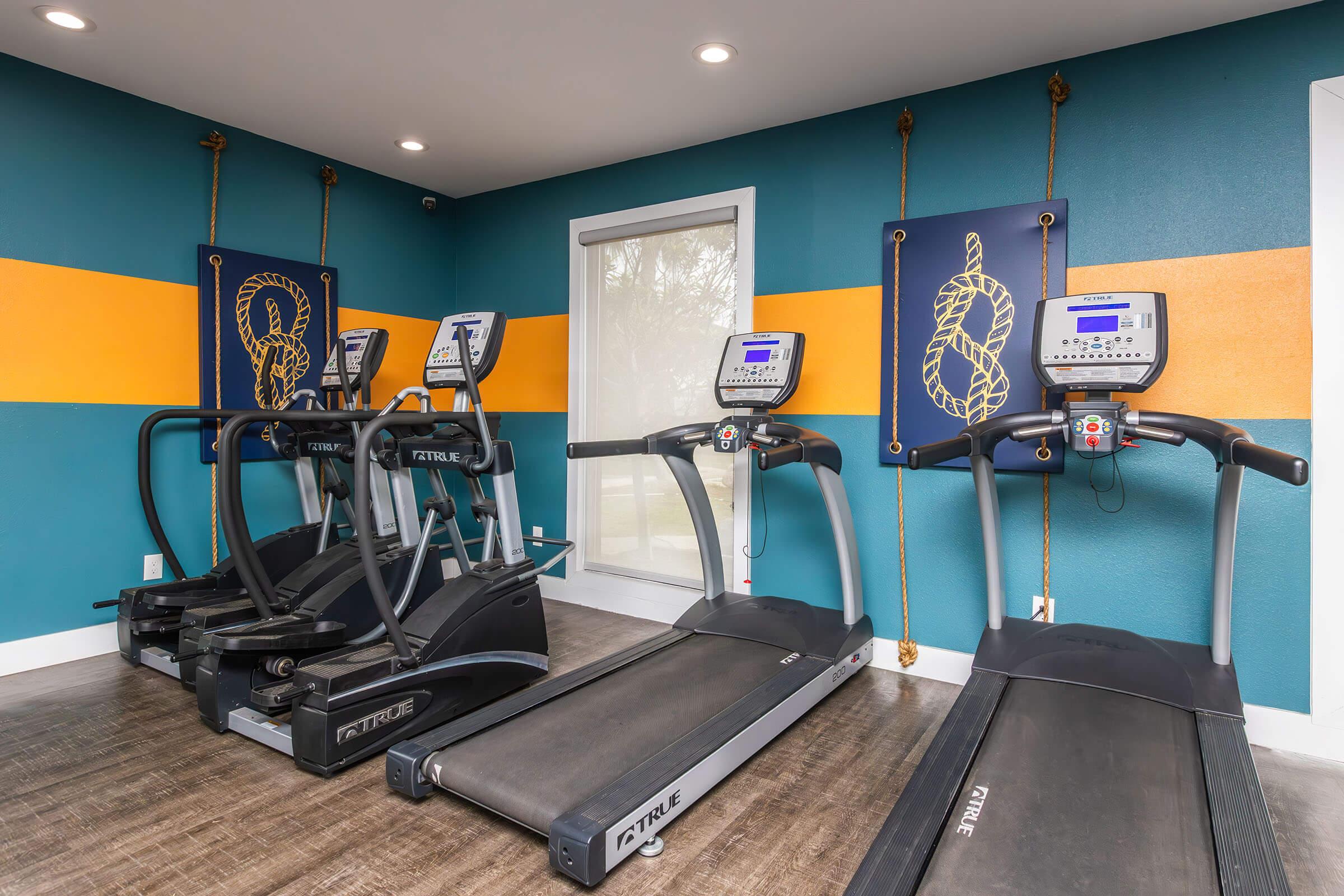 A modern gym with three black treadmills positioned against a vibrant teal wall featuring orange stripes and decorative wall art. The area is well-lit with natural light coming through large windows, creating a welcoming and energetic workout space.